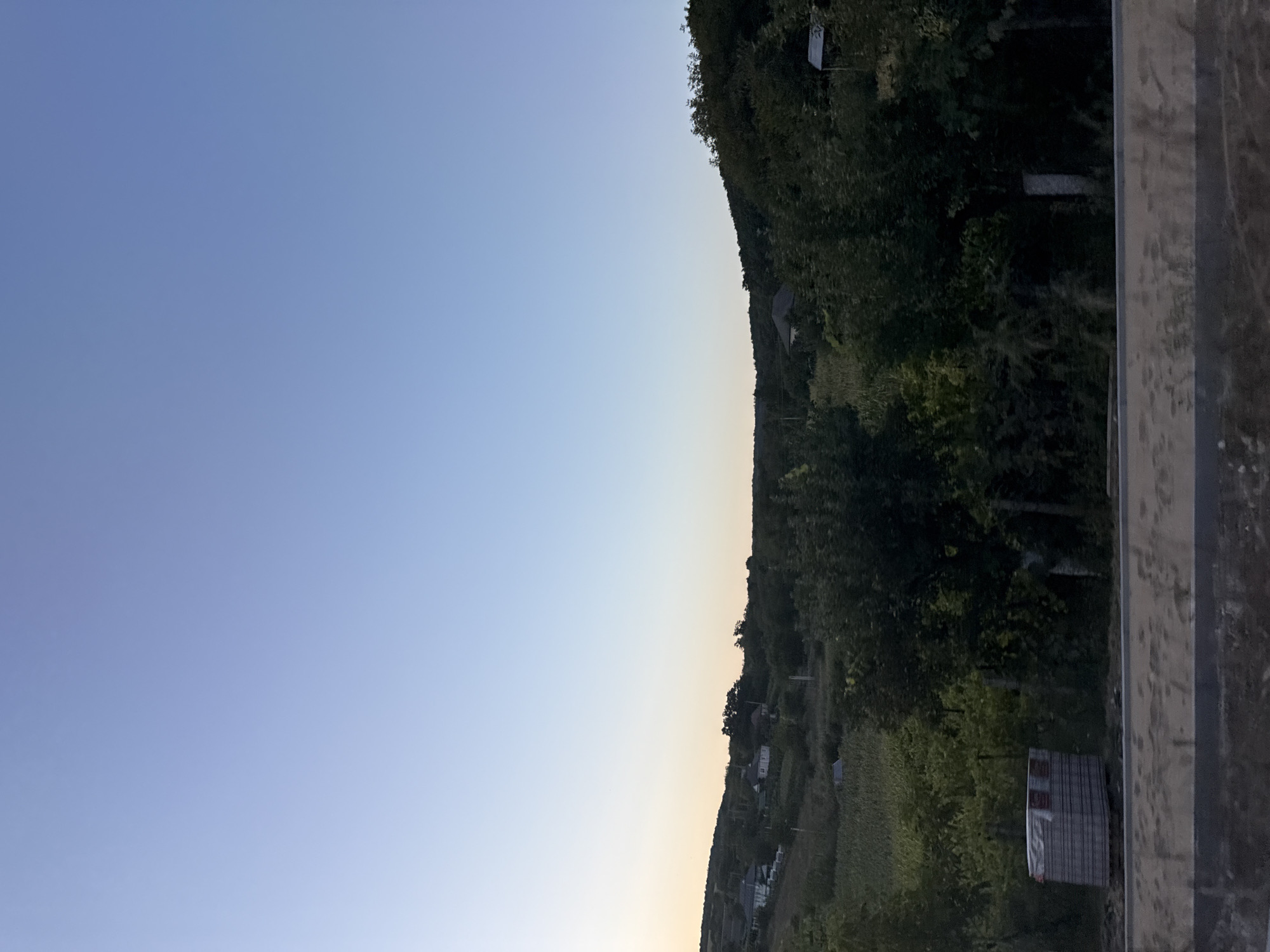 The image shows a rural landscape at dusk in Sărata-Mereșeni, Moldova. The sky is a clear, gradient blue, transitioning to a pale yellow at the horizon. The terrain is hilly and covered with lush vegetation, including trees and what appears to be farmland. Several houses with red roofs are nestled among the trees on the slopes. In the foreground, there is a stack of materials on a pallet, possibly for construction, and a dirt road or path. The scene is peaceful and natural.