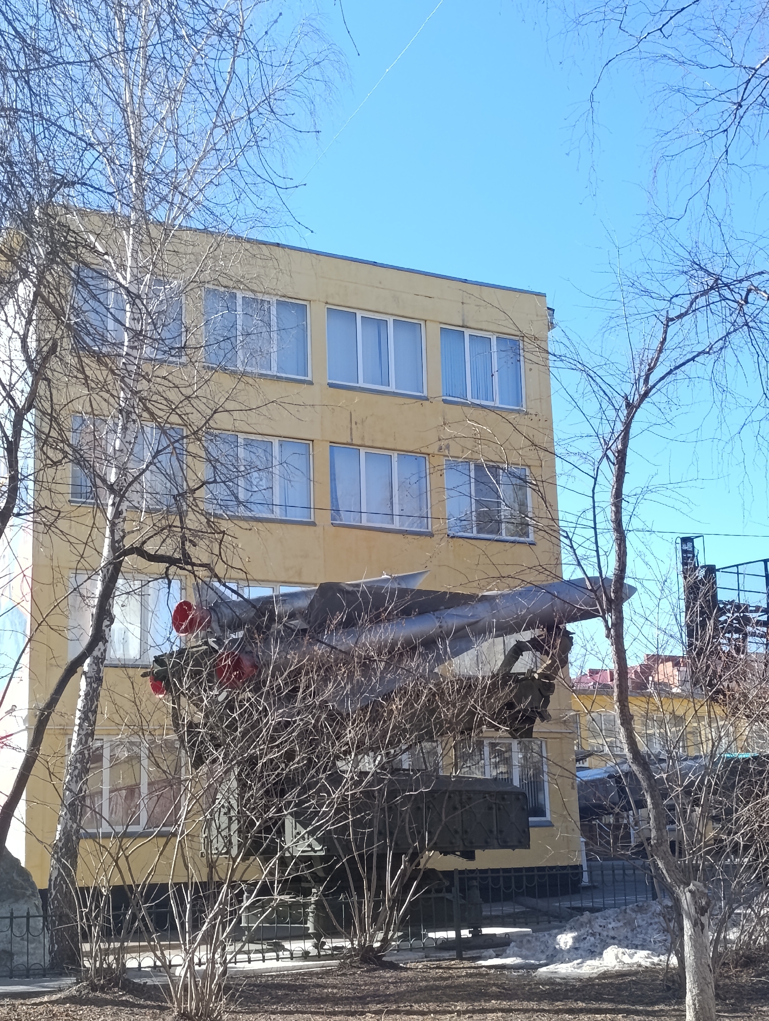 In Krasnoyarsk, Russia, a four-story yellow building stands behind a bare tree and an anti-aircraft missile launcher. The launcher is partially obscured by bushes. Remnants of snow are visible on the ground.