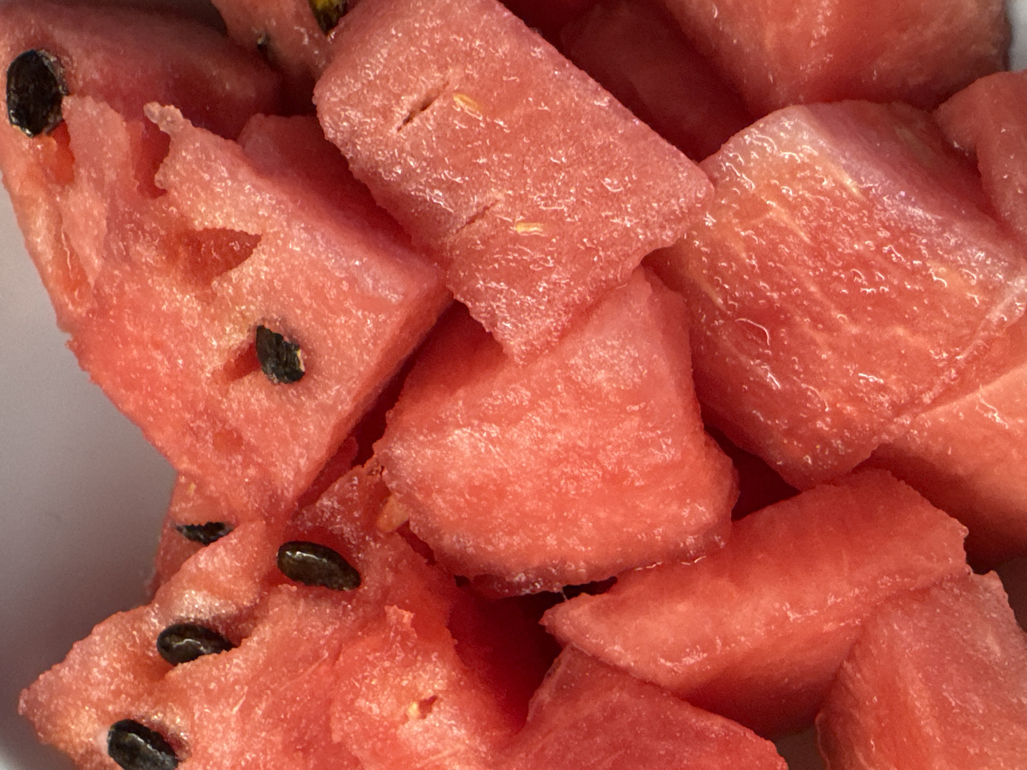 This is a close-up, overhead shot of chopped watermelon. The bright pink flesh of the watermelon is visible, with some pieces showing black seeds and others appearing seedless. The pieces are irregularly shaped, suggesting they were hand-cut. The lighting is bright, highlighting the glistening texture of the fruit. There is no visible text, people, or setting beyond the watermelon itself. The image focuses solely on the texture and color of the fruit.