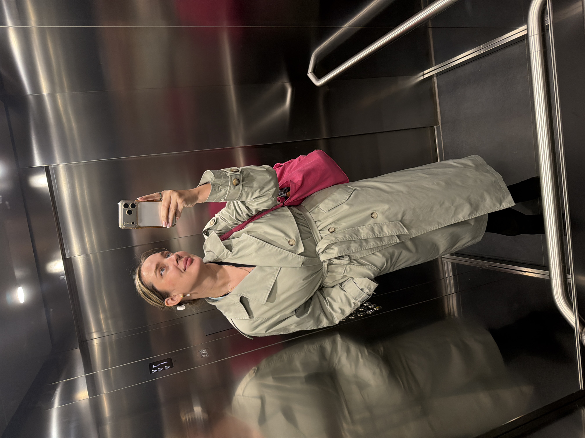A woman in a light-colored trench coat and black pants is taking a selfie with a smartphone in a modern elevator with stainless steel walls and a black floor. She has a bright pink bag slung over her shoulder. The elevator has horizontal handrails and digital floor indicators. The scene may be located in Moscow, Russia.