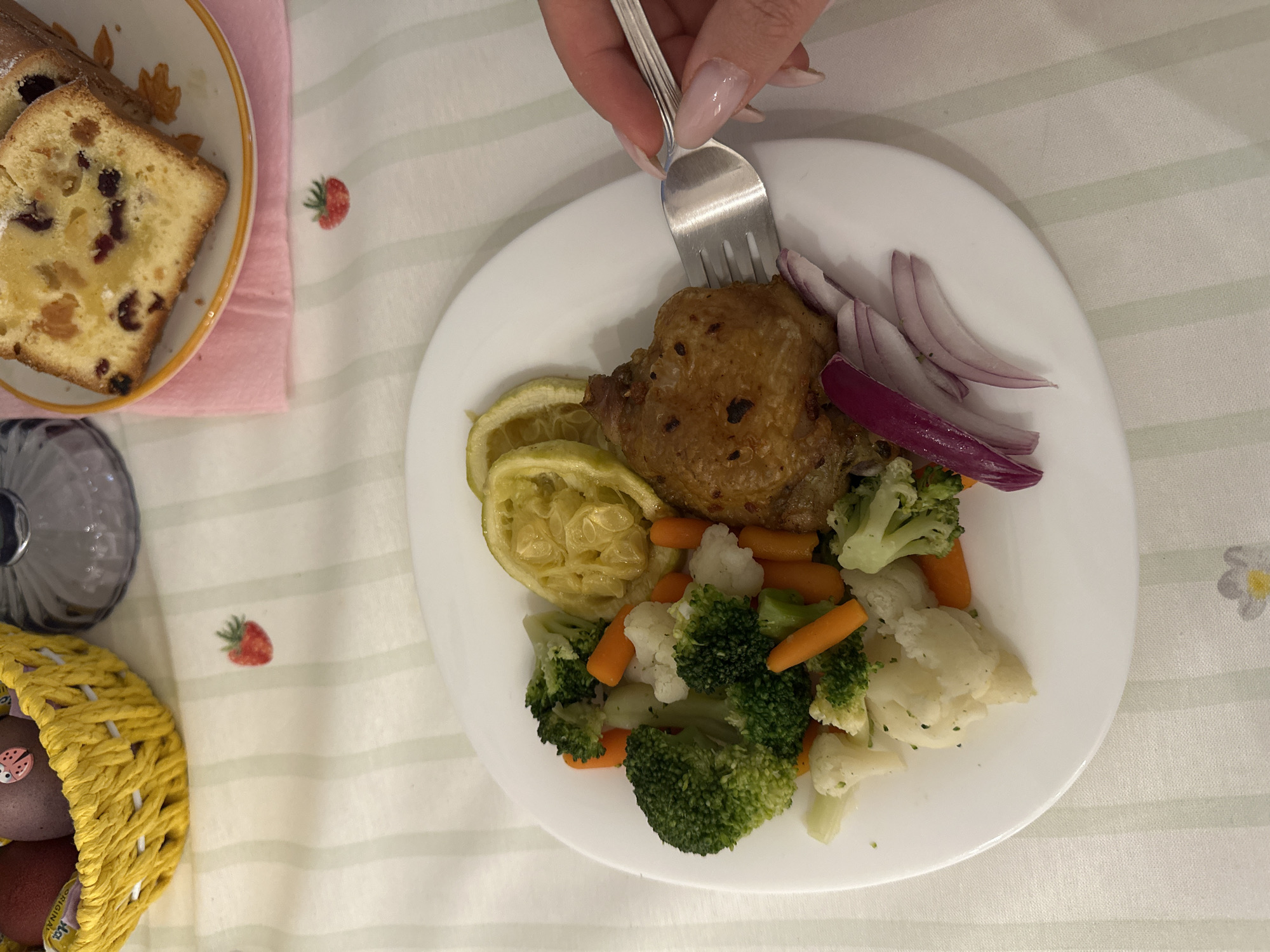 A top-down shot reveals a dining scene. A woman's hand holds a fork, poised over a white plate containing a piece of browned meat, sliced lemon, sliced red onion, and mixed steamed vegetables (broccoli, cauliflower, and carrots). Next to the plate is a slice of fruitcake. The scene rests on a striped tablecloth with strawberry and daisy patterns. A yellow woven basket with Easter eggs sits in the lower left corner, along with a glass vase. The location is Bar, Montenegro, determined by the "La Strada" confectionery sticker on one of the Easter eggs.