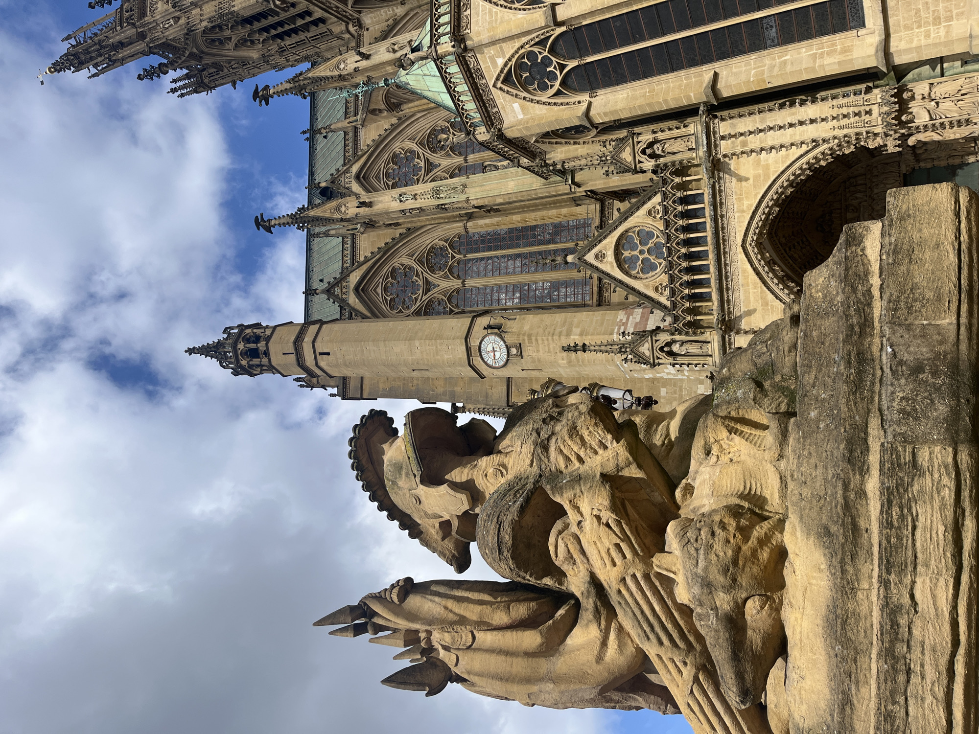 This image shows a low-angle view of Metz Cathedral in Metz, France. In the foreground is a weathered, carved sandstone statue of a man in armor, facing left. The cathedral rises behind the statue, revealing elaborate Gothic architectural details, including pointed arches, intricate tracery, and stained-glass windows. A clock face is visible on one of the towers. The sky is partly cloudy, with patches of blue visible.