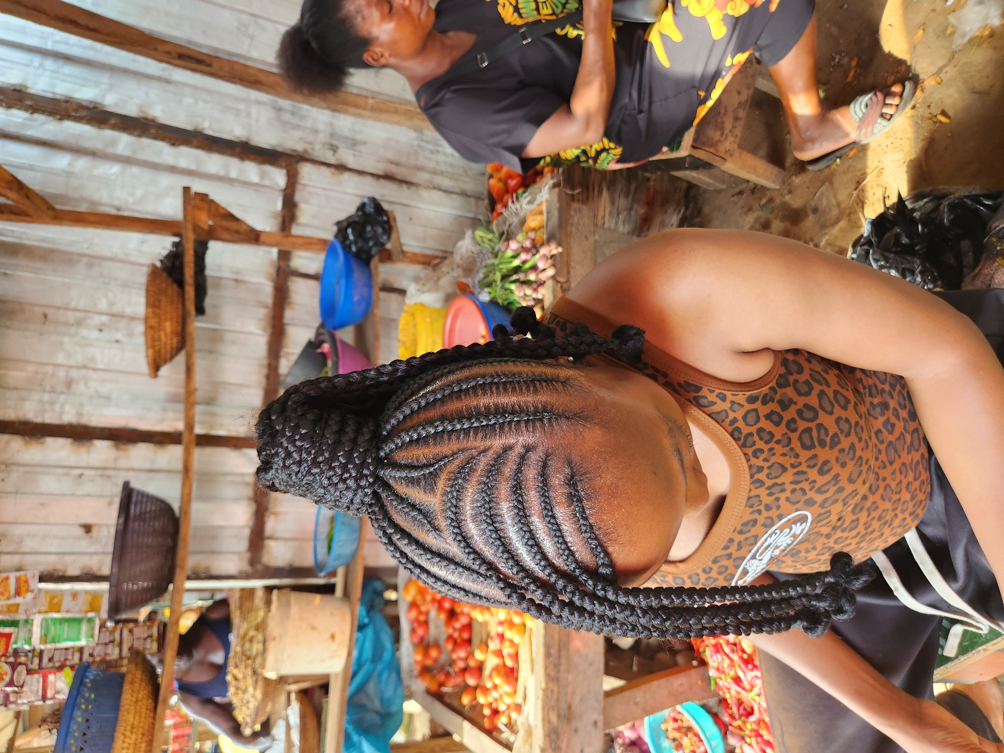 The image depicts an outdoor market scene in Sabon Tasha, Nigeria. In the foreground, a woman with braided hair is partially visible, wearing a leopard-print tank top and dark pants. Her braided hair is styled with a high bun and long braids that hang down. To the right, another woman is seated, wearing a black t-shirt and a colorful patterned skirt, and is engaged with something in her lap. Behind them, market stalls are set up with various goods, including a large display of red tomatoes on a wooden table, bunches of green onions, and other produce in bowls and baskets. Shelves above the stall hold more items, including bags of goods and more baskets. The environment appears rustic, with wooden structures and possibly a dirt or concrete floor. The overall impression is of a busy market environment.