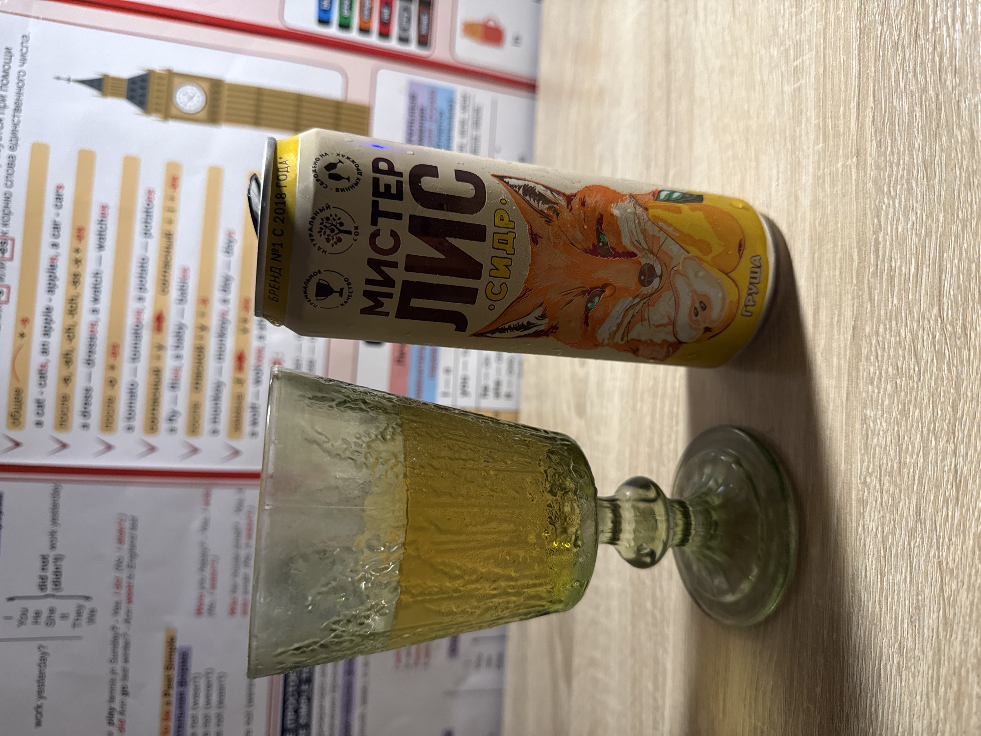 An eye-level, close-up shot captures a glass of pale yellow liquid next to a can of cider. The glass, with its textured surface and wide base, holds a beverage that is lighter in color than the can's label. The can itself is adorned with a cartoon fox and text in Russian, indicating it is a pear cider. In the background, educational materials featuring English grammar exercises and a partial image of Big Ben suggest a learning environment. The scene is set on a wooden surface, implying it is likely indoors, possibly in a home or a study area. The lighting is somewhat dim, with a slight warm cast. There is no indication of the city of Cheboksary, Russia.