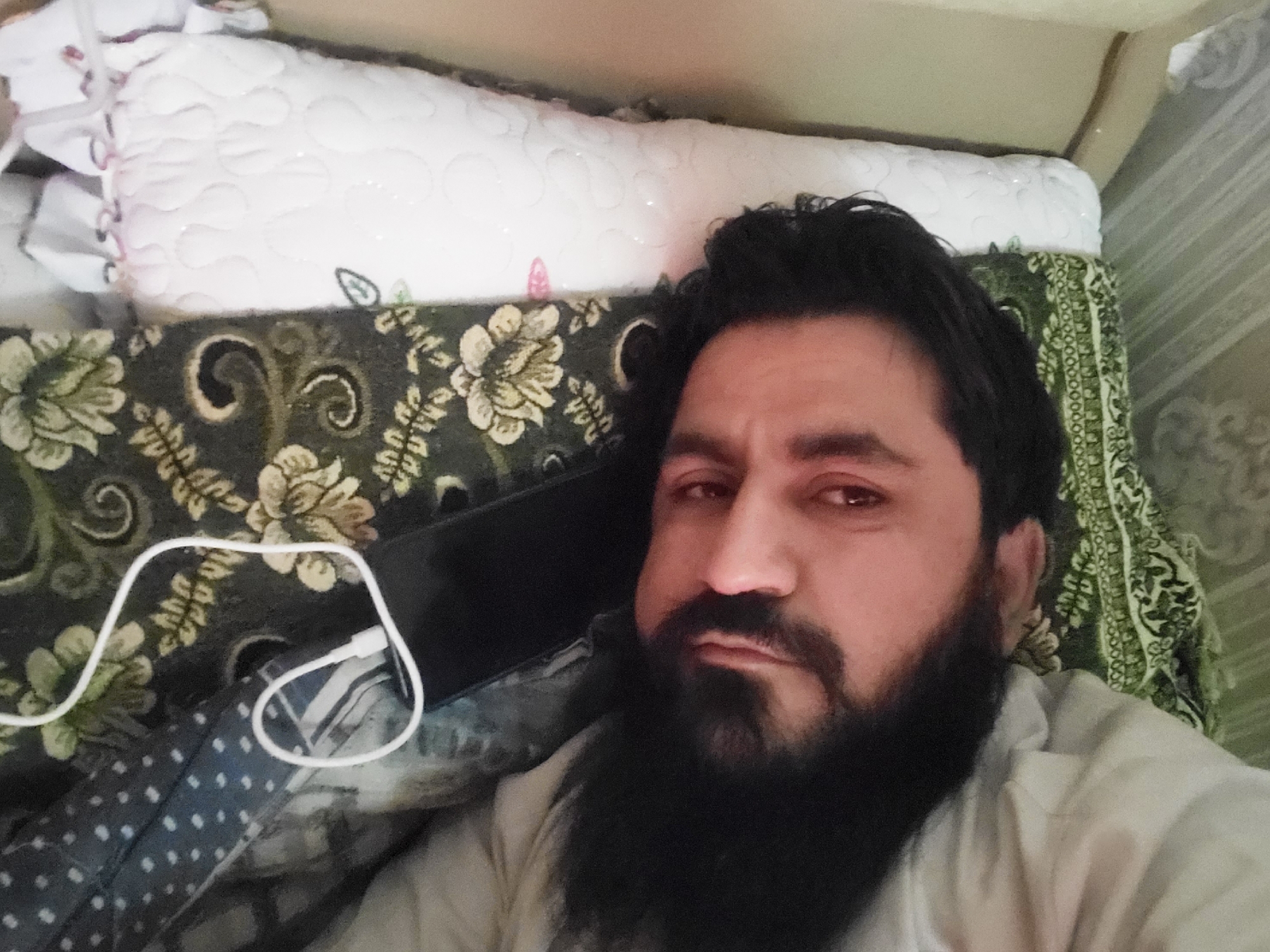 A man with dark hair and a full beard is lying down, taking a selfie. He is positioned against a patterned cushion, with a white, quilted bolster behind him. A white charging cable is visible near the cushion. The location is likely inside a building in Quetta City Tehsil, Pakistan, given the prompt.