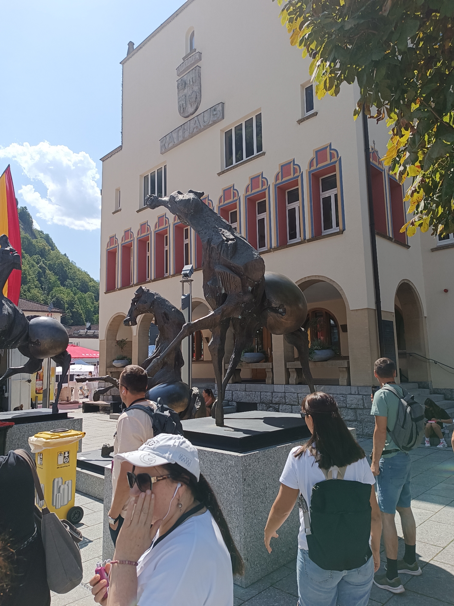 In front of the Rathaus (City Hall) in Vaduz, Liechtenstein, a bronze sculpture depicting abstract horses is prominently displayed. The Rathaus is a light-colored building with arched entrances and windows. Above the main entrance, an emblem is visible, and the word "RATHAUS" is inscribed. A flag, red and yellow, is partially visible on the left side of the image. Several people are present in the scene. In the foreground, a woman wearing a white cap, sunglasses, and a white t-shirt is holding a pink object and touching her face. To her right, another woman in a white t-shirt and blue jeans with a dark backpack is walking away from the camera. A man in a light-colored t-shirt and blue shorts with a backpack is standing to the right, facing the Rathaus. Other individuals and elements like a yellow trash can and potted plants are also discernible. The environment appears to be a public square with a paved ground. The sky is clear with some clouds, and trees are visible in the background.