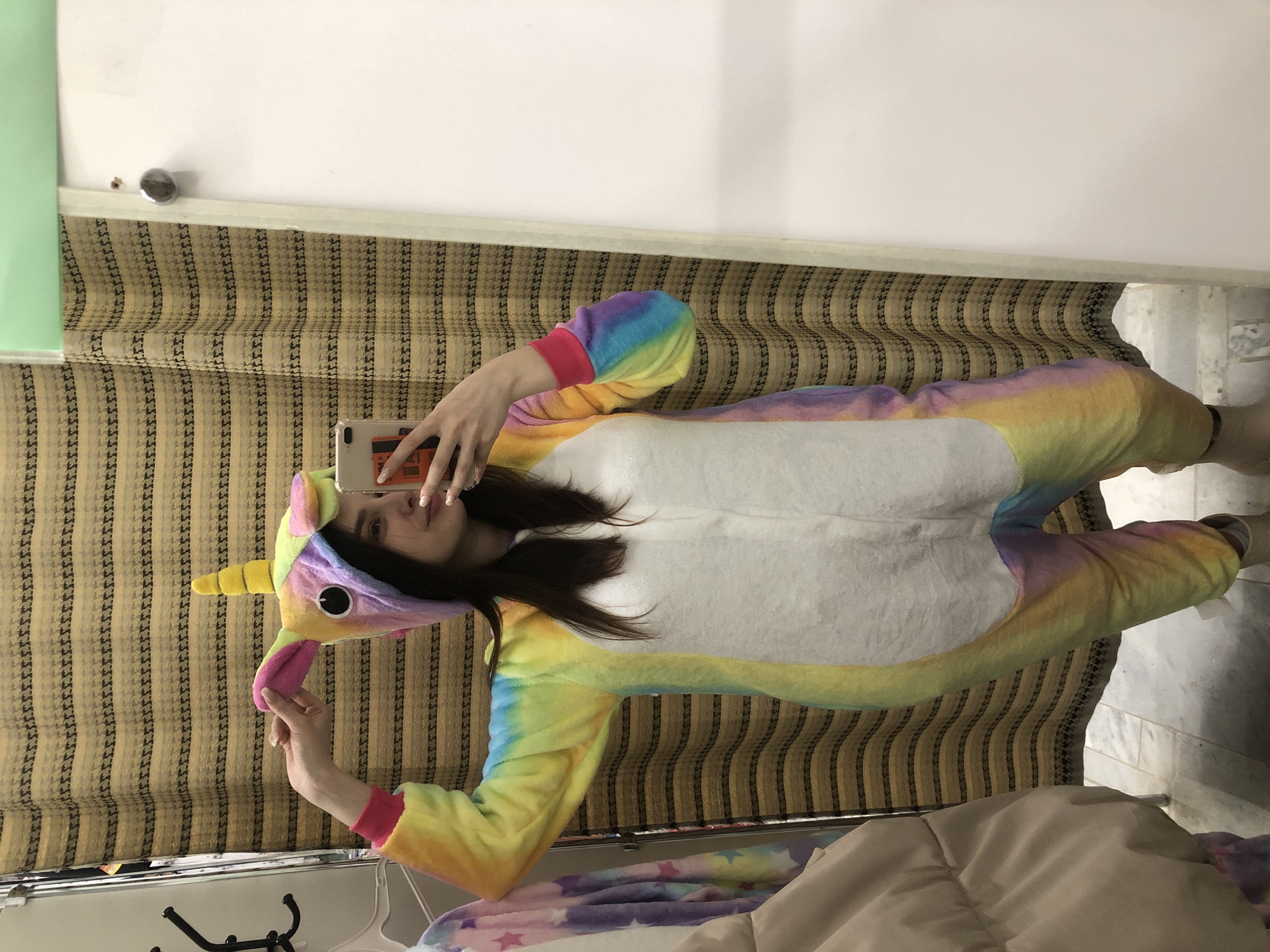 A person wearing a unicorn onesie is taking a selfie in a room. The person is holding a smartphone in their right hand. The room has a light-colored wall and a window with a beige and brown patterned shade. The person is wearing a colorful unicorn onesie with a white torso and a yellow horn on the hood. A partial view of bedding is in the bottom of the frame. The location is not identifiable as Chaikovsky, Russia.