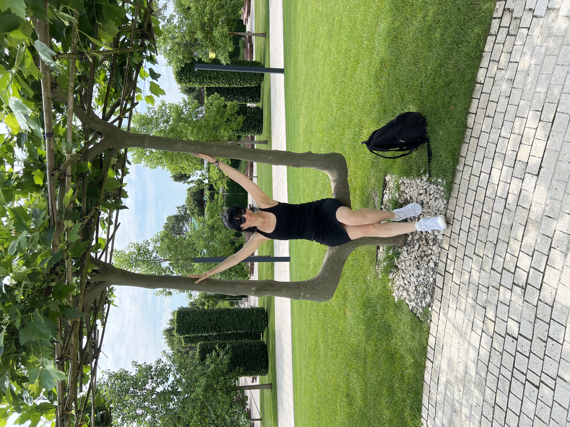 A woman is seated on a uniquely shaped tree in a park. The tree branches are trained to form a seat with two upright trunks. The woman is wearing a black tank dress, white sneakers, and sunglasses. She has her arms raised, resting on the tree's branches. A black backpack is placed on the grass next to her. The park setting features a manicured lawn, paved walkways, and neatly trimmed hedges. In the background, there are more trees, benches, and a lamppost. The sky is partly cloudy. This scene is located in Park "Krasnodar" (also known as "Galitsky Park") in Krasnodar, Russia.