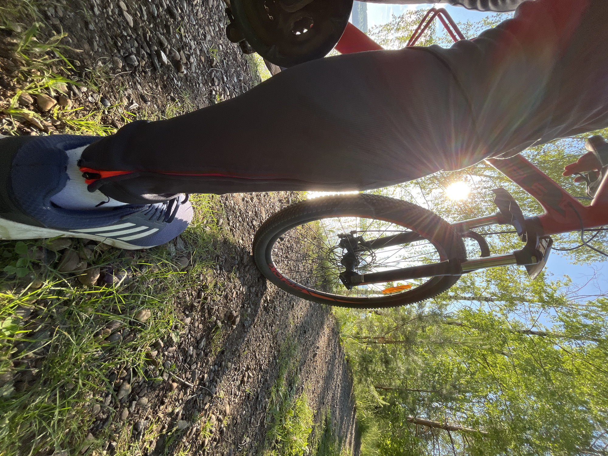The image depicts a close-up, low-angle view of a person's legs and a red bicycle standing on a gravel path next to a body of water, likely the Kama River. The sun is low in the sky, creating lens flares and casting long shadows. Green trees and foliage surround the scene, suggesting an outdoor, natural environment. The person is wearing dark athletic pants with red accents and blue athletic shoes. The bicycle has a front suspension fork and a partially visible wheel with orange detailing. The ground is covered in small stones and patches of green grass. The overall atmosphere is serene and hints at a recreational activity like cycling. The location is identified as Chaikovsky, Russia, a city situated on the Kama River, known for its picturesque natural surroundings and recreational opportunities.