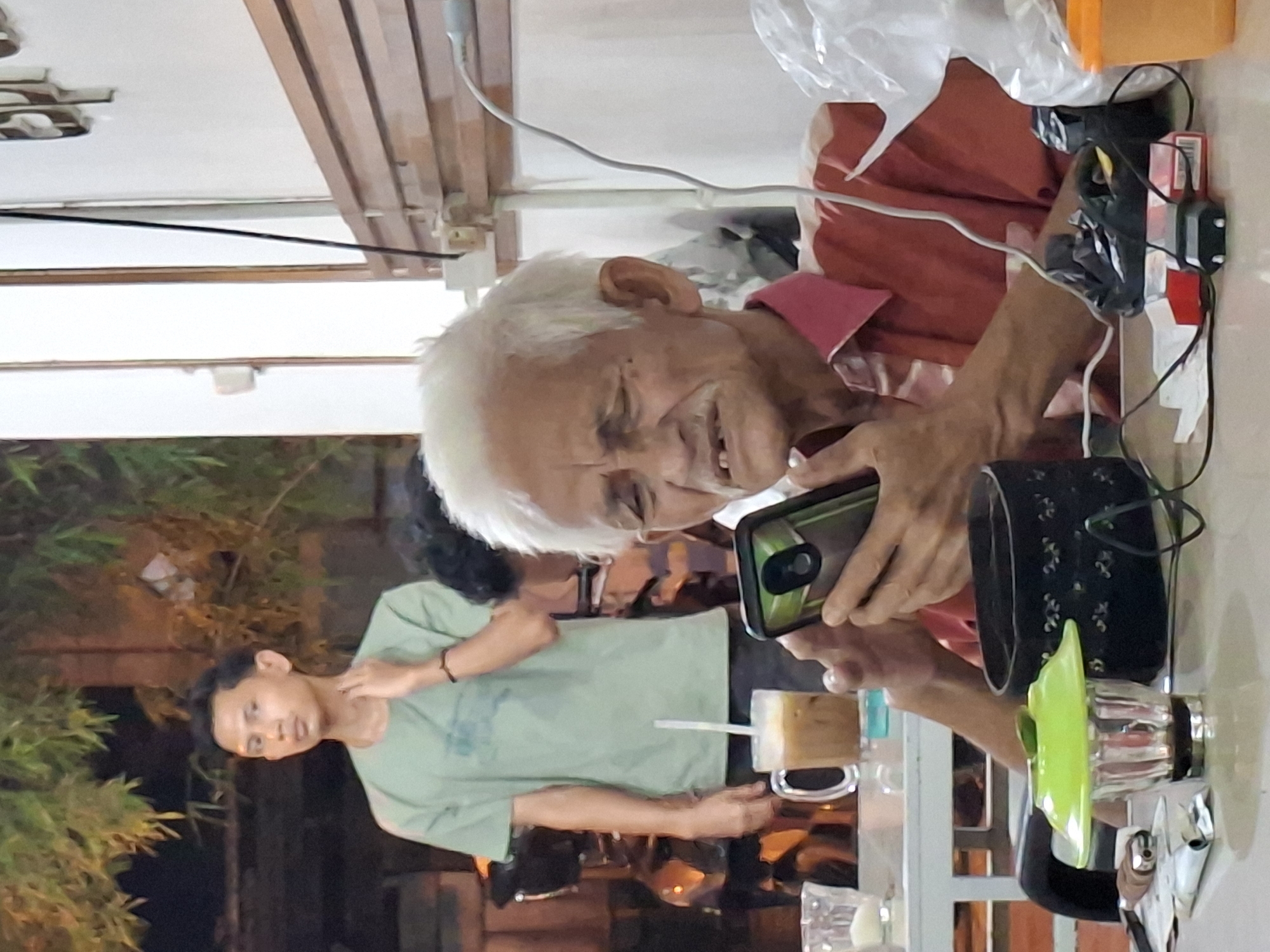 An elderly man with white hair is seated at a table, looking at a smartphone. He wears a red shirt. Beside him is a young man in a green shirt scratching his neck, and both are in what appears to be an outdoor seating area with greenery in the background. On the table are drinks, a mug, and other items. The location is likely a café or restaurant in Lhokseumawe, Indonesia.