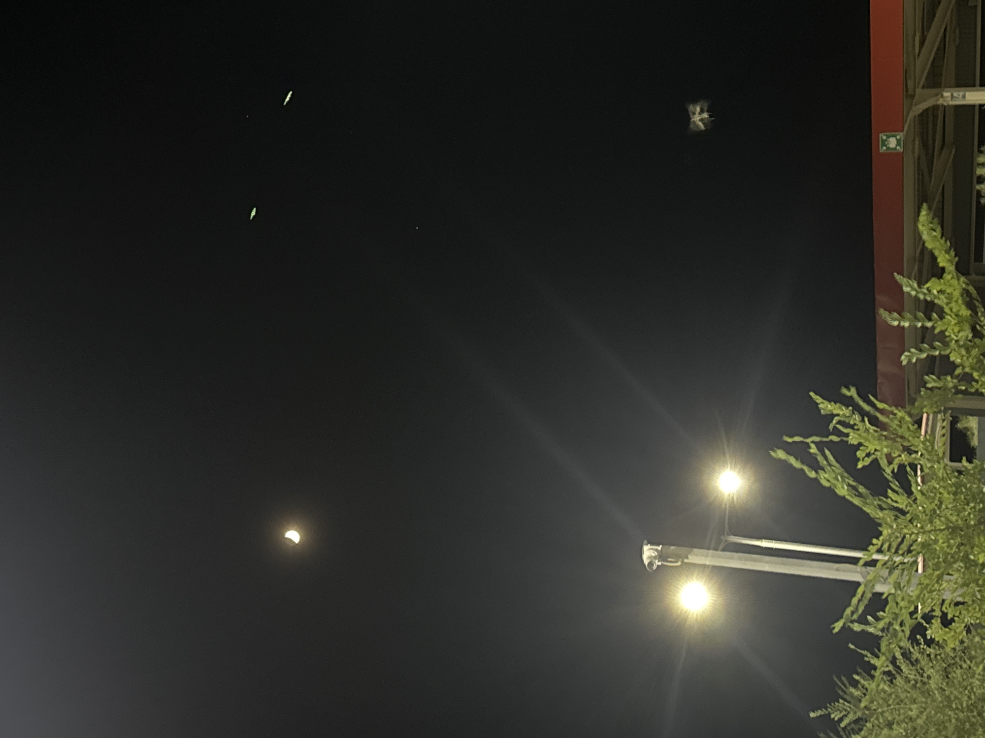 In the night sky above Unknown, Uzbekistan, a crescent moon hangs in the upper left quadrant of the frame. Below it, two streetlights with visible light flares illuminate the scene. A lamppost with a security camera is positioned in the lower center, with two bright light bulbs. In the foreground, green foliage from a tree obscures the bottom part of the image. In the middle right of the sky, a small, blurry object is visible, possibly a drone or a bird. There are also three faint, elongated green lights in the upper right of the sky.
