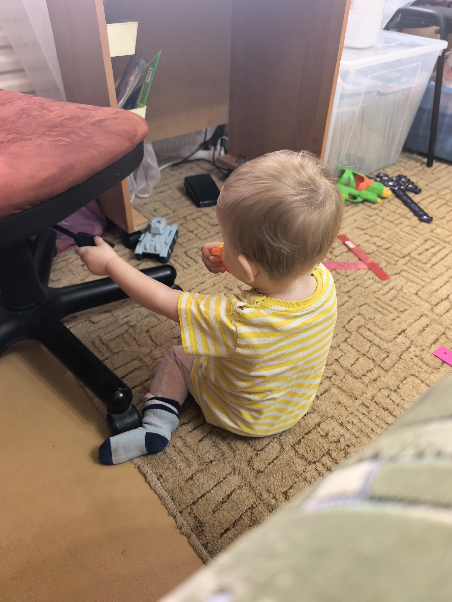 A young child, seen from behind, sits on a patterned brown carpet in an indoor setting, likely a home in Sterlitamak, Russia. The child, with light blonde or light brown hair, is wearing a yellow and white striped t-shirt, pink pants, and blue, white, and dark blue striped socks.
The child is actively engaging with their surroundings, reaching their left hand out to touch or manipulate a lever or control mechanism on the black base of a red or pink upholstered office chair. In their right hand, they hold a small, orange object, which could be a toy or a snack.
The room contains typical household items. To the left, part of a wooden desk or shelf unit is visible, with some documents or papers stored in a small holder underneath. To the right, a large, clear plastic storage container with a white lid stands against the wall. Various toys are scattered across the carpet, including a grey tank, a green water gun, a dark purple toy gun, and several red and pink plastic strips or rulers. Wires and a small black electronic device are also visible on the floor near the desk. The lighting suggests an indoor scene during the day or under artificial light. No discernible text is visible on any objects.