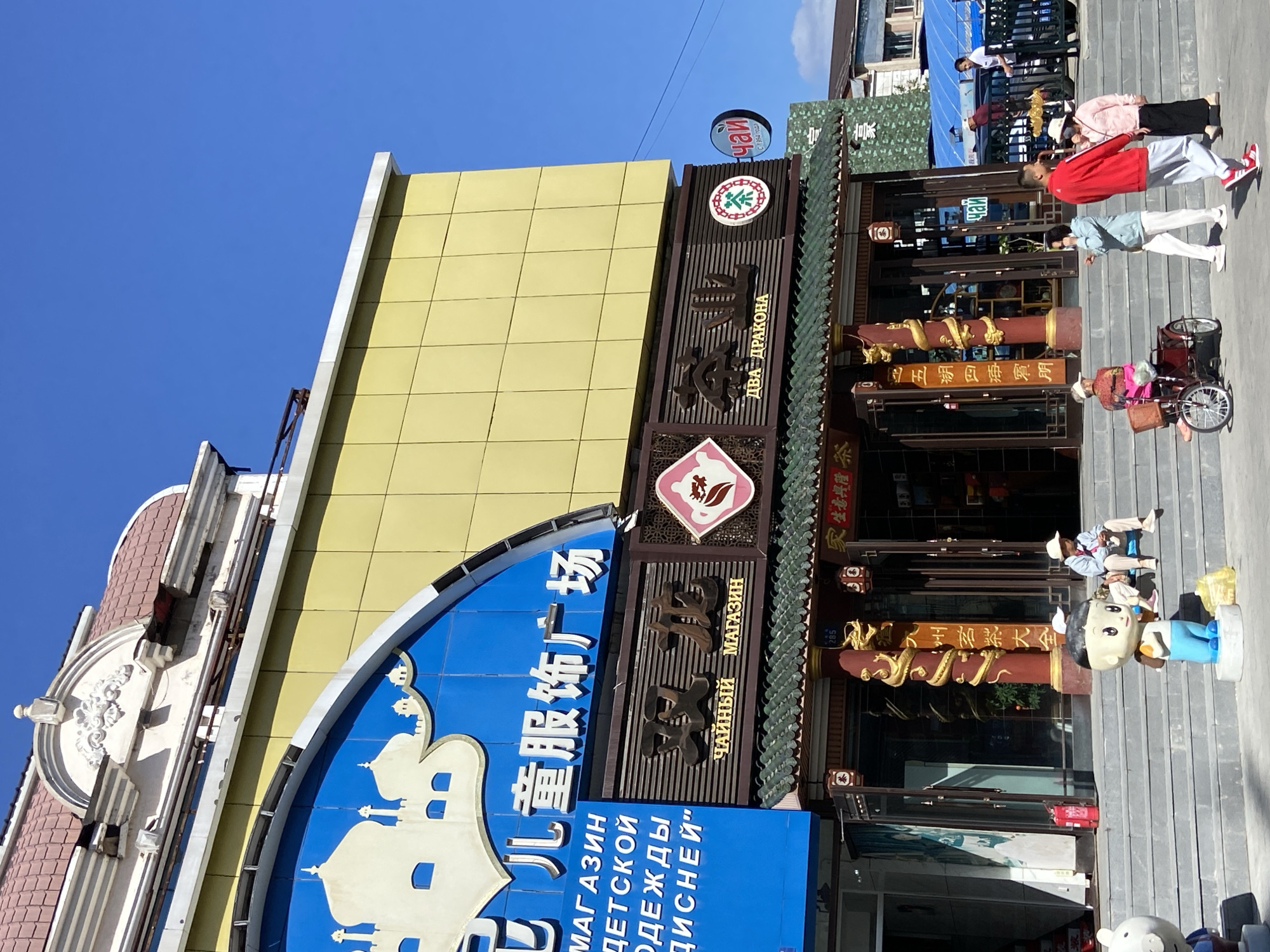 The image depicts a street scene in Xing'an Subdistrict, China, focusing on the exterior of a commercial building. The building features a bright yellow facade on its upper section, contrasted with a blue arched sign above the entrance. This blue sign displays white text and an illustration of white domes, suggestive of a cultural or religious motif. Below this, a darker brown fascia features Chinese characters and smaller signs with text in Russian, indicating a mixed-language environment. One sign reads "ЧАЙНЫЙ МАГАЗИН" (Tea Shop) and another "ДВА ДРАКОНА" (Two Dragons). A circular logo with a green tea leaf design is also visible.
The ground level of the building has large glass windows and doors, with decorative red pillars featuring golden dragon motifs. A traditional green tiled roof extends over the entrance area. In front of the building, a set of wide grey steps leads up to the entrance. Several people are present on and around these steps. To the left, a child-like statue stands on a pedestal. A person wearing a white hat and a light blue jacket is seated on the steps, holding something in their hands. To their right, another individual, also in a white hat, sits on the steps. Further to the right, a person rides a red tricycle. A group of people are walking up the steps, including a man in a red jacket and light blue pants, a woman in a light blue jacket and white pants, and a woman in a pink jacket and black pants.
The environment appears to be a sunny day with a clear blue sky. In the background, other buildings and structures are visible, including a black metal fence and some blue tarpaulins. The overall impression is a busy commercial area with a blend of cultural influences, as indicated by the signage.