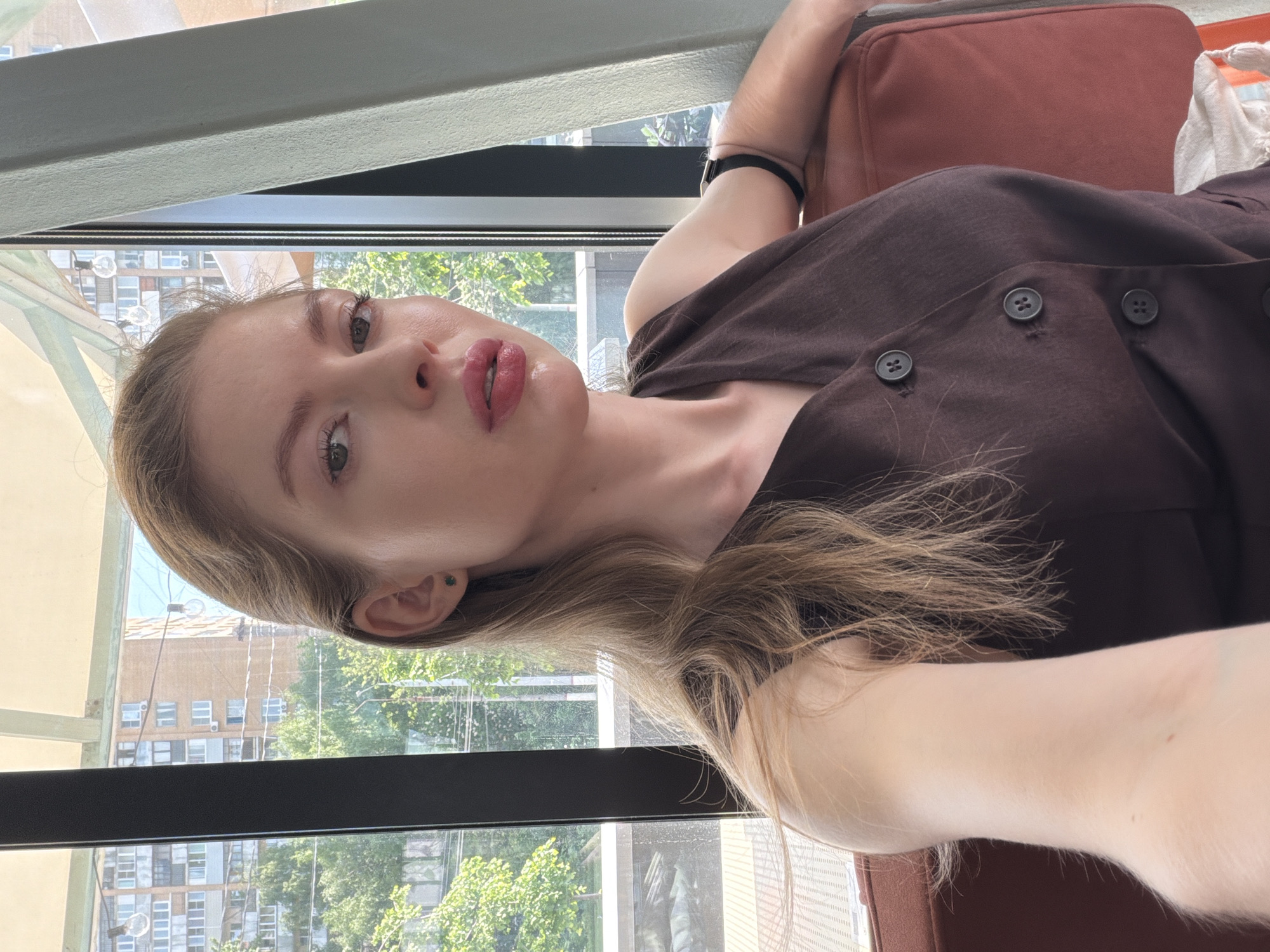 A fair-skinned woman with light brown hair is shown in a selfie taken indoors. She is wearing a brown button-down shirt. The background shows a cityscape with apartment buildings and greenery, visible through a window. Based on visual clues, the buildings outside the window could be located in Khimki, Russia, based on the style of architecture.