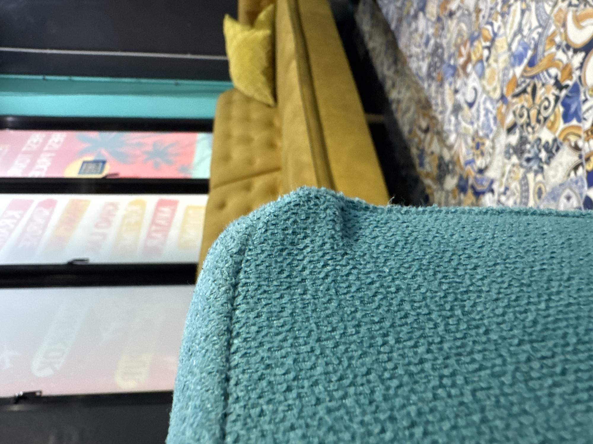 This media file features a close-up shot of the textured fabric of a turquoise sofa cushion, angled upwards. Behind the cushion, a yellow tufted sofa with a small yellow pillow is visible. Further back, the image shows a patterned fabric with a blue, gold, and white ornate design. The scene appears to be indoors. In the background, there are reflections or signs with text, including what looks like palm tree graphics and lettering in pink and yellow. The lighting suggests it is daytime, possibly indoors with artificial light or natural light filtering in. The overall impression is one of interior decor.