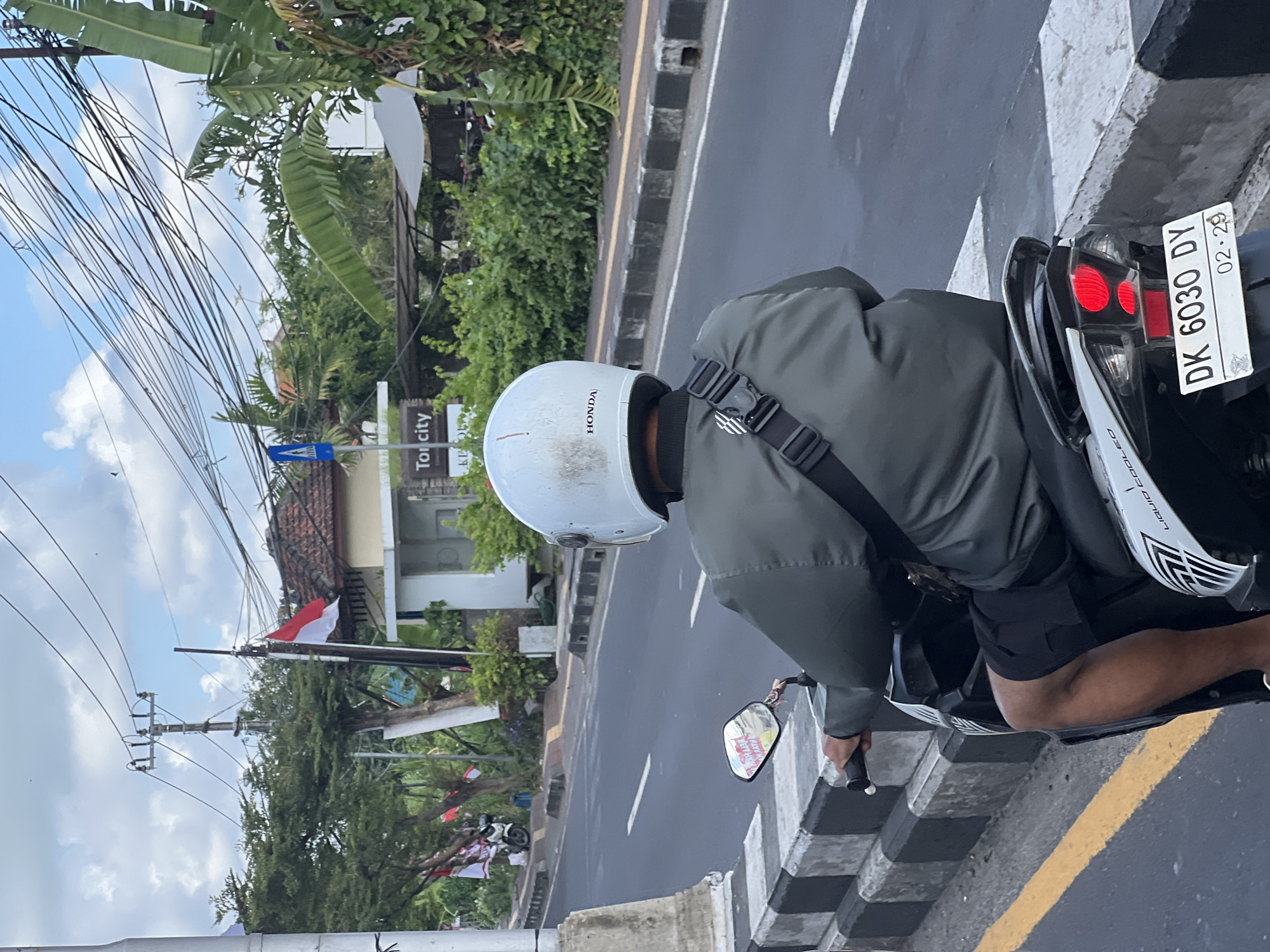 A person wearing a white helmet and a dark green jacket rides a scooter on a street in Denpasar, Indonesia. The scooter has a license plate that reads "DK 6030 DY" and "02.29". In the background, there are trees, power lines, and buildings. One building has a sign that says "Tor City". Indonesian flags are visible on poles in the background. The street has a yellow line on the left side and a checkered curb. The sky is cloudy.