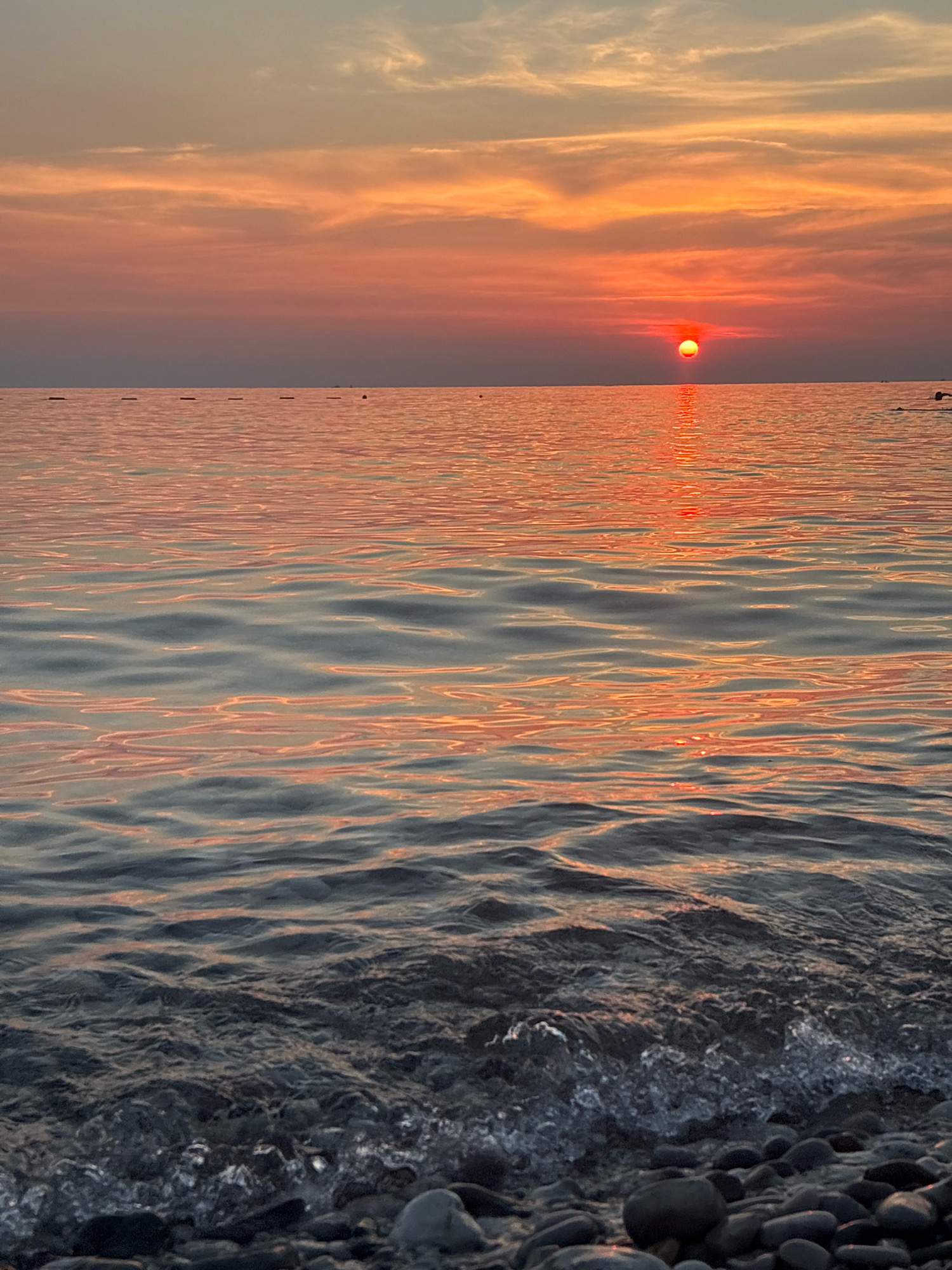 A serene sunset over the Adriatic Sea at Bar, Montenegro. The sun, a vibrant orange orb, is just above the horizon, casting a warm glow across the sky and reflecting on the gentle ripples of the water. The sky transitions from a soft peach and pink near the sun to a pale, dusky blue at the top. In the foreground, dark, smooth pebbles line the shore, with small waves lapping at them, creating a foamy white edge. The water is a mix of deep blues and grays, interspersed with the shimmering orange and pink reflections of the sunset. A few buoys are visible in the distance, marking a safe swimming area.