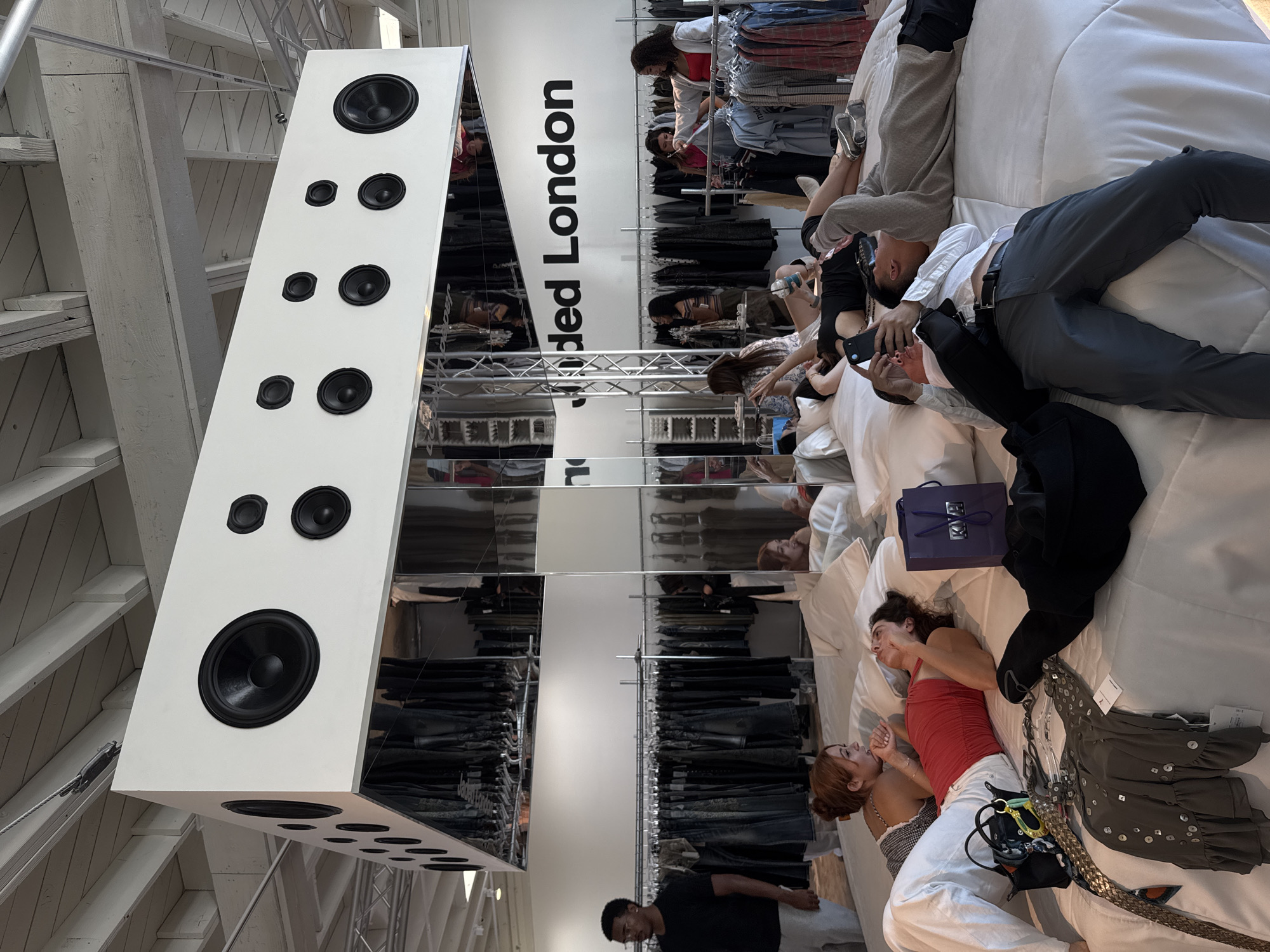 A high-angle shot depicts an interior space in West Hollywood, California, United States of America. The scene features several individuals in a retail environment. The individuals are lying on a white bed or display area. Some are using phones, while others are resting or conversing. Clothing racks filled with garments are visible in the background, as well as large signage with the inscription "Jaded London". Suspended from the ceiling is a large, white rectangular speaker, which is reflected in a mirrored surface. Overhead beams and structural elements of the building are also visible. A purple gift bag is visible near the individuals.