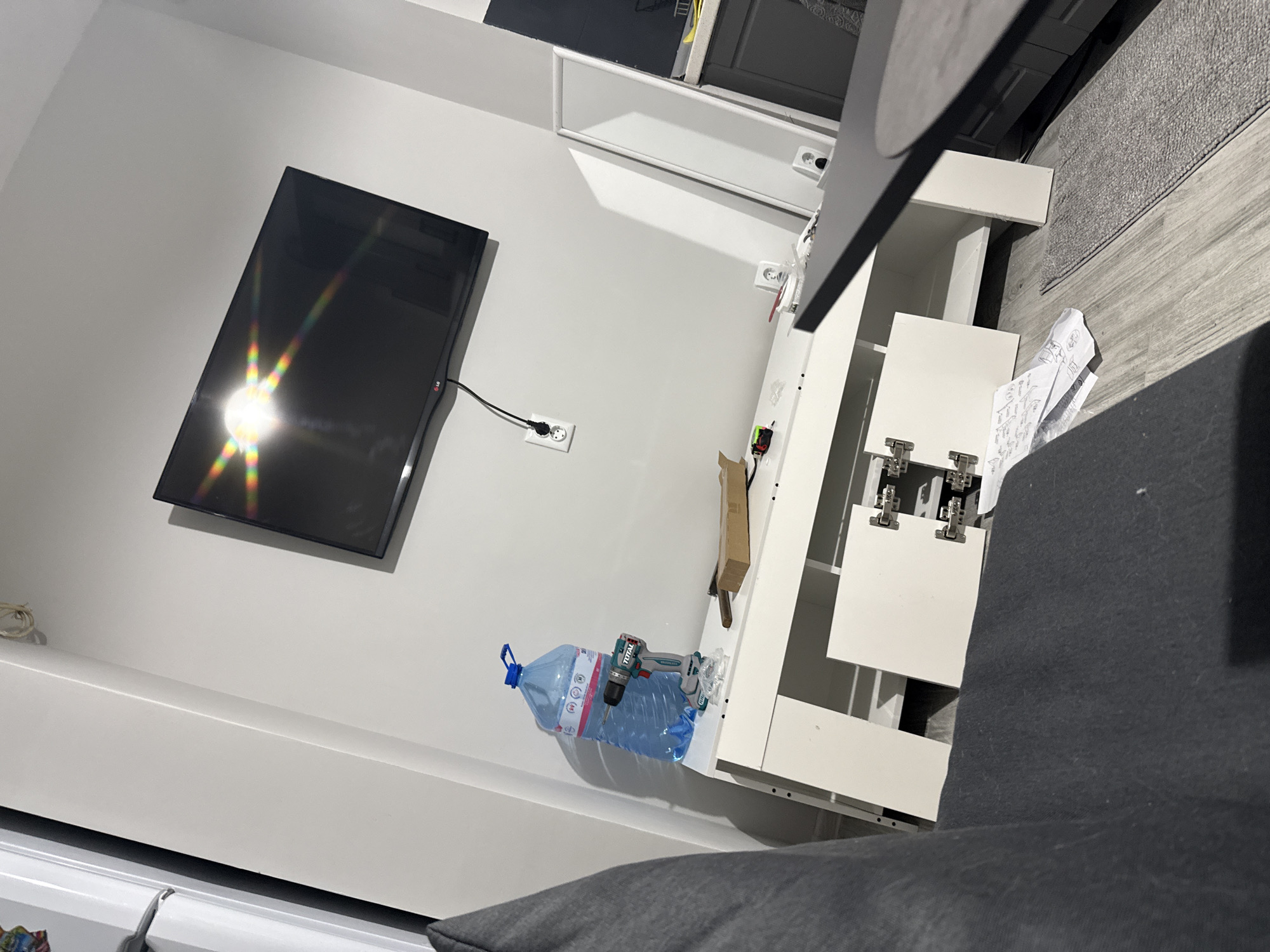 A low-angle, indoor shot shows a living space. On the left, a flat-screen TV is mounted on a white wall. An electrical outlet is visible near the TV. A white cabinet or shelving unit is to the right. A large water bottle and a power drill rest on one of the cabinet shelves. A piece of furniture, possibly a sofa, occupies the bottom center of the frame, obscuring the lower portion of the cabinet. The floor is visible in the upper right corner and appears to be made of wood or laminate. The location is Stara Zagora, Bulgaria. No people are visible in the image.