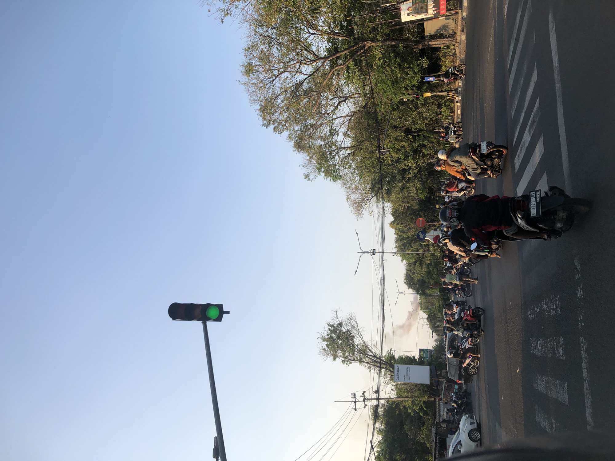 The image depicts a street intersection in Gelam, Indonesia, under a clear blue sky. A traffic light, currently showing green, hangs overhead, controlling the flow of traffic. A variety of vehicles, predominantly motorcycles, are present at the intersection, some waiting, while others are in motion. Pedestrians are not clearly visible, but the presence of a crosswalk indicates their potential use of the area. Trees line the sides of the street, providing a natural element to the urban landscape. Smoke can be seen rising in the background, suggesting a possible incident or industrial activity in the distance. The overall scene captures a typical day at a bustling intersection in Indonesia.