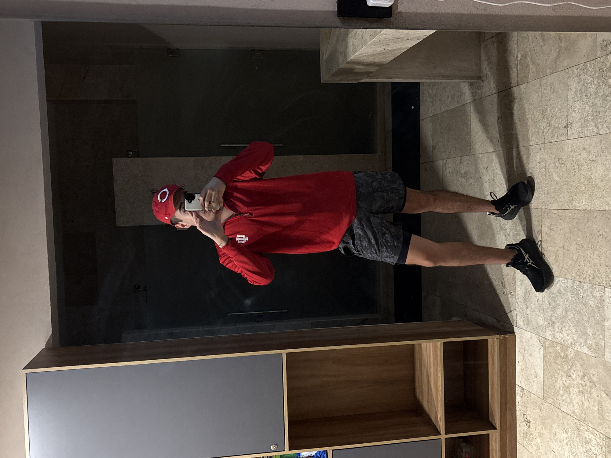 A person is seen taking a mirror selfie in an indoor setting in San Bernardino Tlaxcalancingo, Mexico. The individual, likely male, is standing upright, holding a silver/white smartphone to capture their reflection. They are wearing a red long-sleeved shirt with a white logo resembling a stylized "IU" or trident on the left chest, and a red baseball cap featuring a white "C" logo. Their attire also includes dark, patterned athletic shorts and black athletic shoes with white accents, possibly bearing an Asics logo.
The setting reflected in the mirror appears to be a modern room, possibly a bathroom or dressing area, with a light-colored tiled floor and dark, reflective surfaces in the background, which might be glass shower doors or dark walls. A light-colored counter or shelf is also visible in the reflection. Surrounding the mirror, elements of the actual room include a light-colored wall and a modern cabinet or shelving unit at the bottom, featuring both gray drawer/door fronts and open wooden cubbies. The lighting is artificial, casting subtle shadows on the tiled floor.