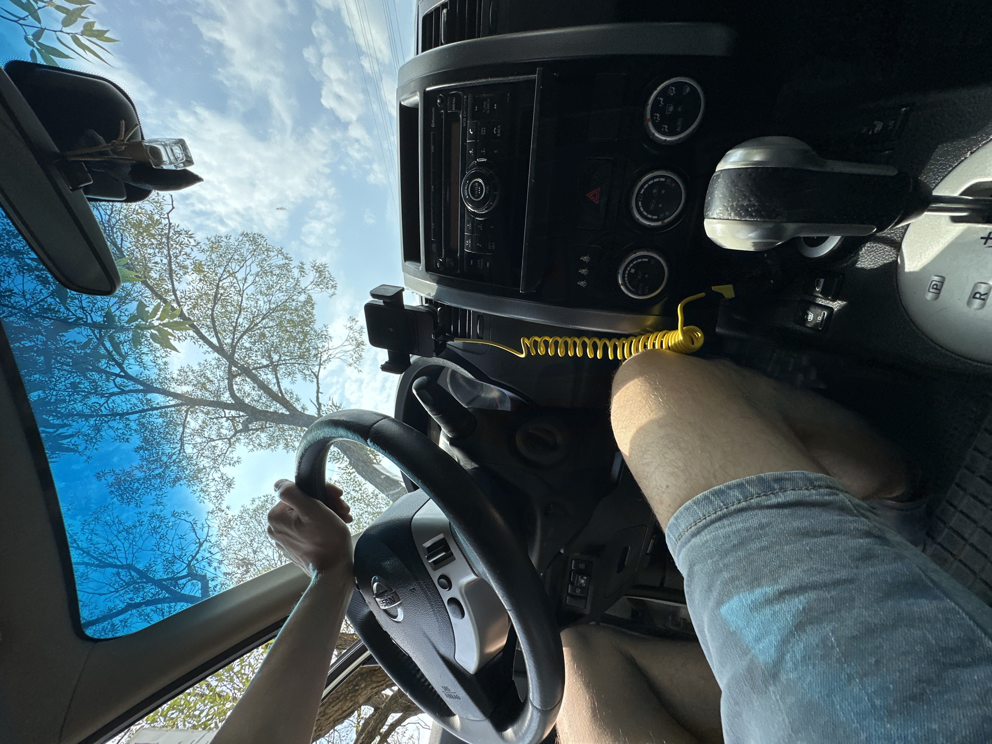 The image shows the interior of a car from the perspective of the driver. The driver's hands are on the steering wheel. The dashboard with the radio and air conditioning controls is visible. A yellow coiled cord with a phone holder is attached to the dashboard. The windshield shows trees and a blue sky with clouds. The car appears to be parked on a street in Makiv, Ukraine, given the presence of trees and the architectural style of the surroundings, although this cannot be definitively confirmed.