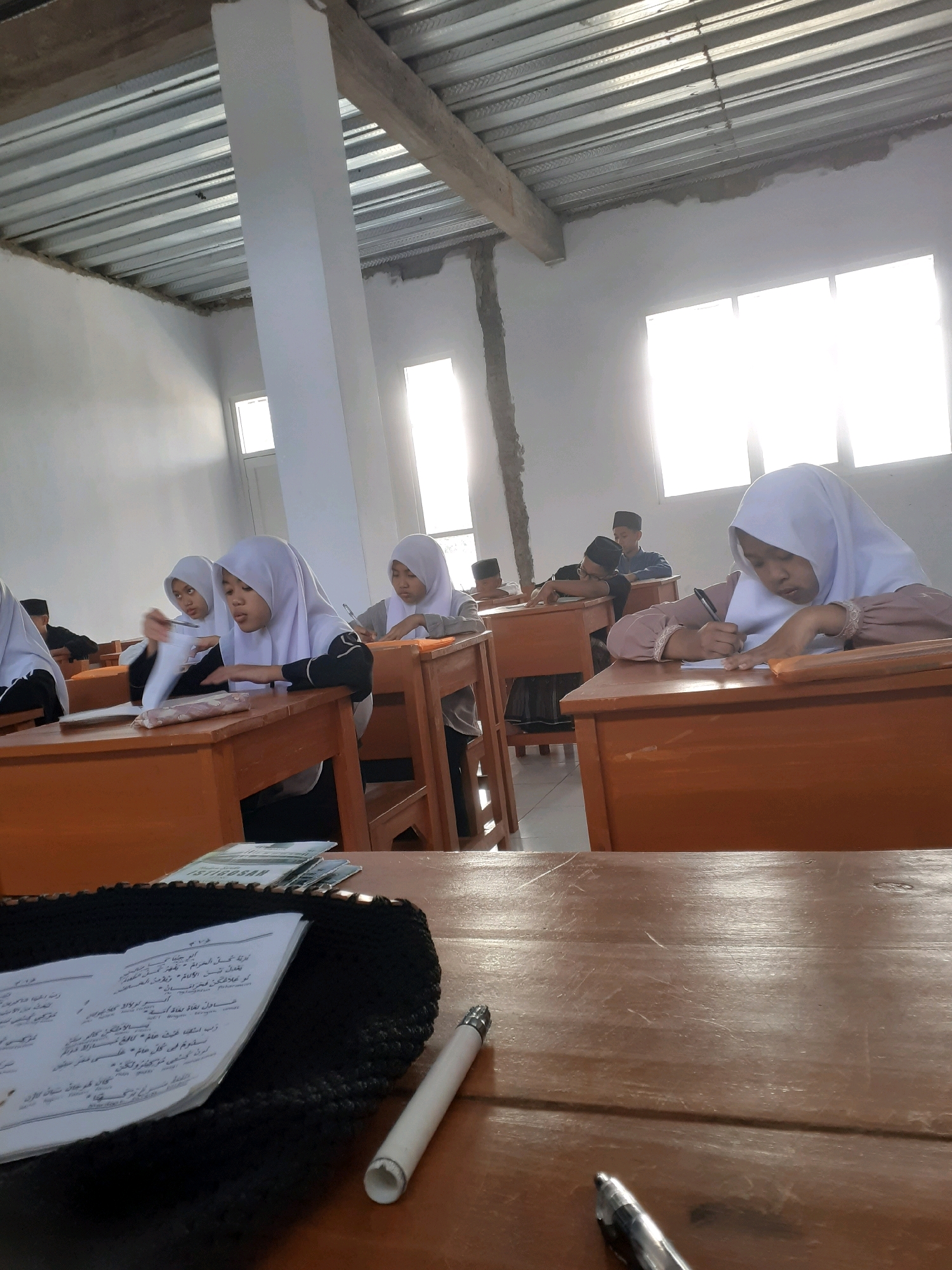 The image depicts an indoor scene, likely a classroom, with several young female students wearing hijabs seated at wooden desks. The room appears to be under construction or newly built, as indicated by the exposed ceiling with corrugated metal and wooden beams, and unfinished walls. Sunlight streams in from windows on the right side of the frame, creating bright highlights. The students are engaged in activities such as writing and reading. One student in the foreground is actively writing on a piece of paper with a pen, while others are looking at their books or papers. The desks are simple, wooden structures. In the foreground, a stack of papers with Arabic script and a white pen are visible on a desk. The overall impression is one of a modest educational setting.