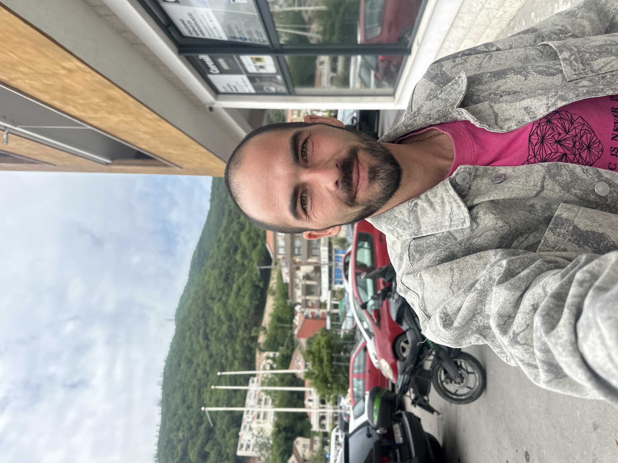 A selfie of a man with a beard, wearing a pink t-shirt and a gray camouflage jacket. The picture appears to be taken outdoors, in the city of Budva, Montenegro. In the background, there's a building with windows and a mountainous landscape. Cars and motorcycles are parked in the street. The sky is partially visible, indicating daytime.