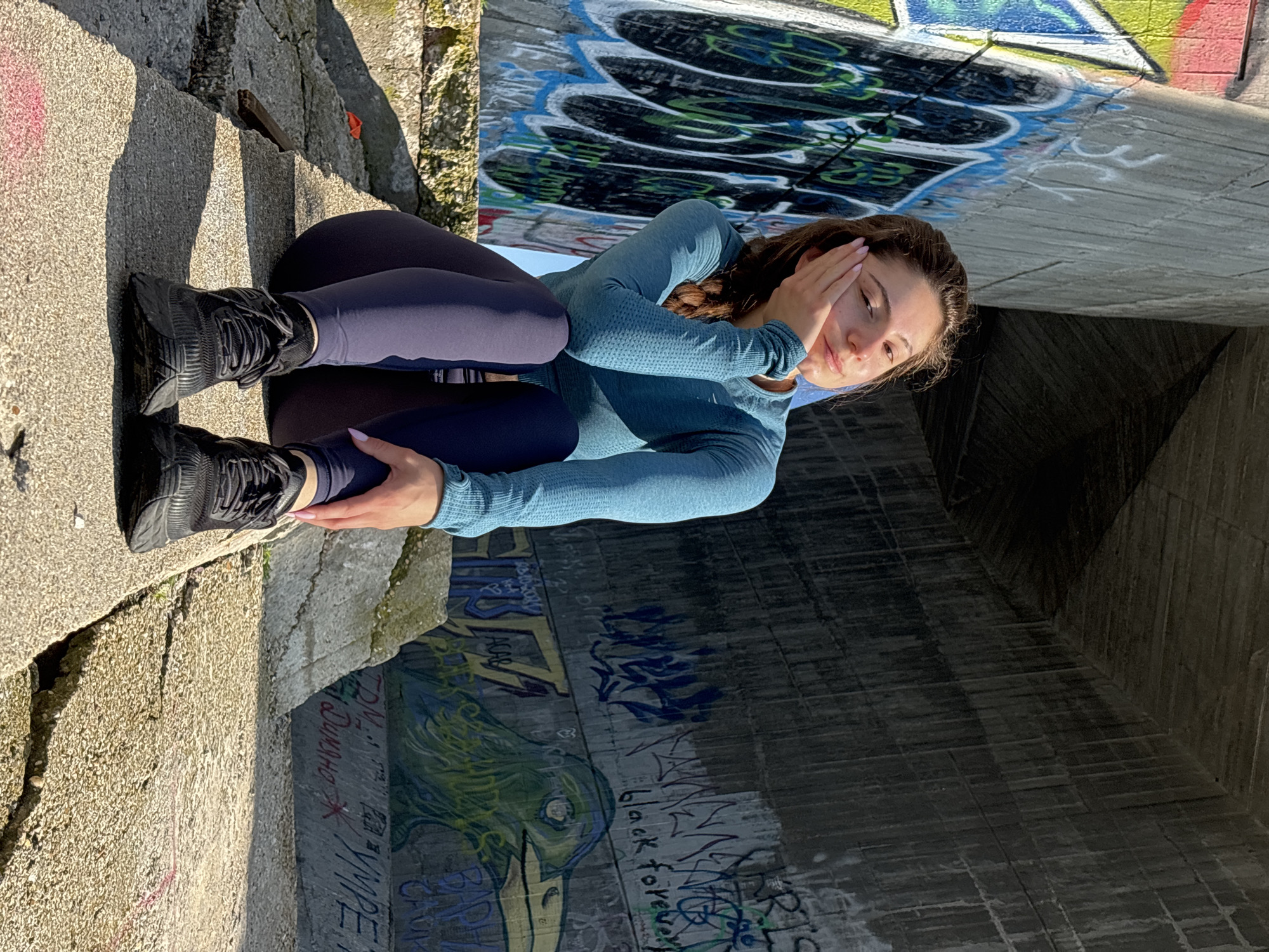 A young woman is seated on rough concrete steps under a large concrete structure, likely an underpass or bridge. She has long brown hair, styled in a braid over her left shoulder, and is wearing a light blue long-sleeved top, dark blue leggings, and black athletic shoes. Her fingernails are painted light pink. She is sitting with her knees bent, and her right hand is raised to shield her eyes from bright sunlight, which casts strong shadows and illuminates her face. Her expression appears calm or subtly smiling.
The surrounding concrete walls and ceiling are extensively covered in vibrant graffiti, featuring various colors including blue, green, yellow, red, black, and white. Visible text within the graffiti includes "black forever," along with partial words like "DUMANA" and "YPPE." The setting suggests an urban environment, and the harsh shadows indicate a bright, sunny day, possibly morning or late afternoon.