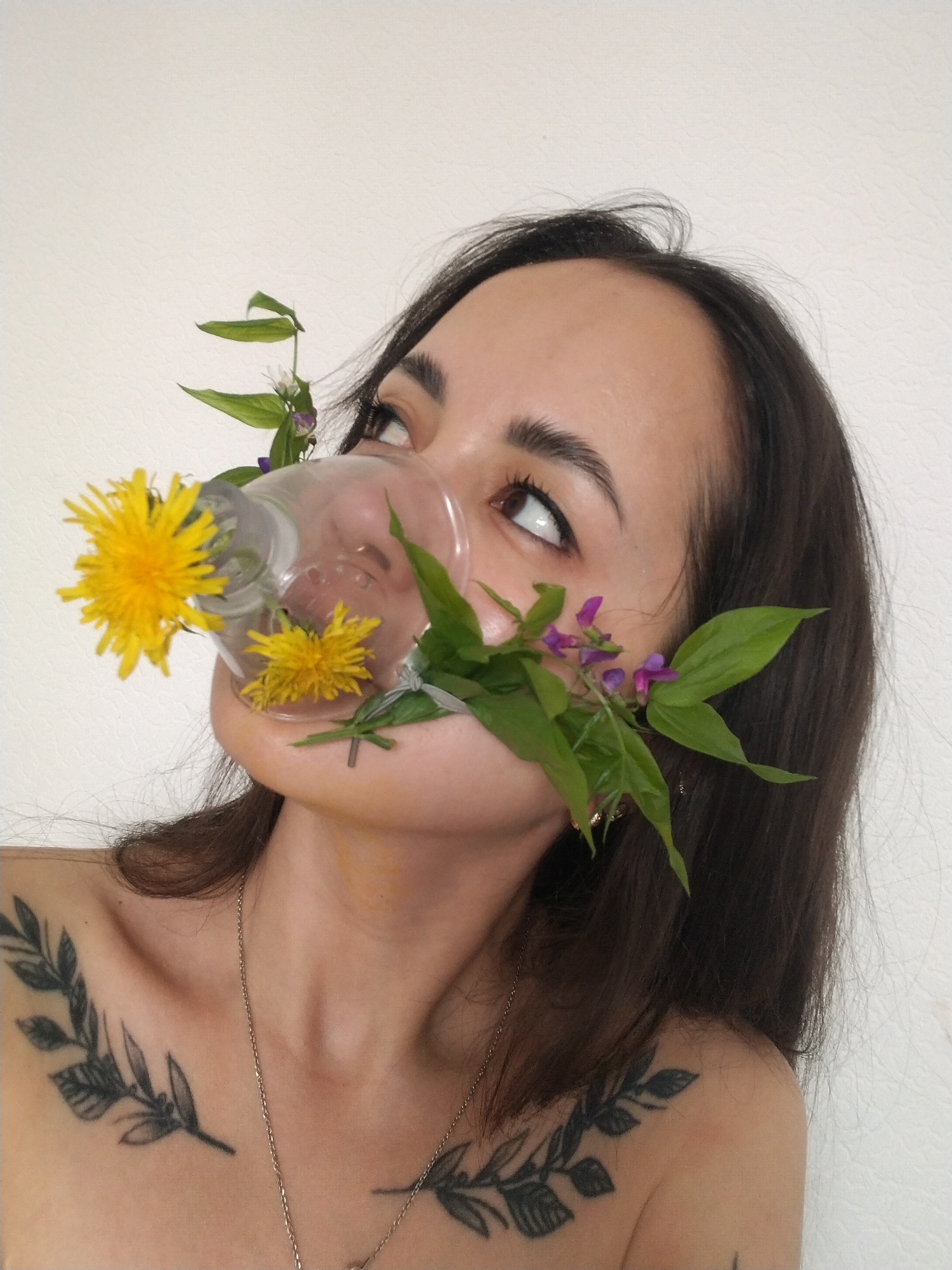 A woman is depicted in an eye-level portrait. She wears a glass object over her mouth and nose, which is adorned with dandelions and small purple flowers with green leaves. Her eyes are upturned to the upper right of the image. The woman has dark hair and a fair complexion. Two black leaf tattoos are visible on her upper chest. She wears a silver necklace. The background is a plain white wall. This image was taken in the city of Noviy, Russia.