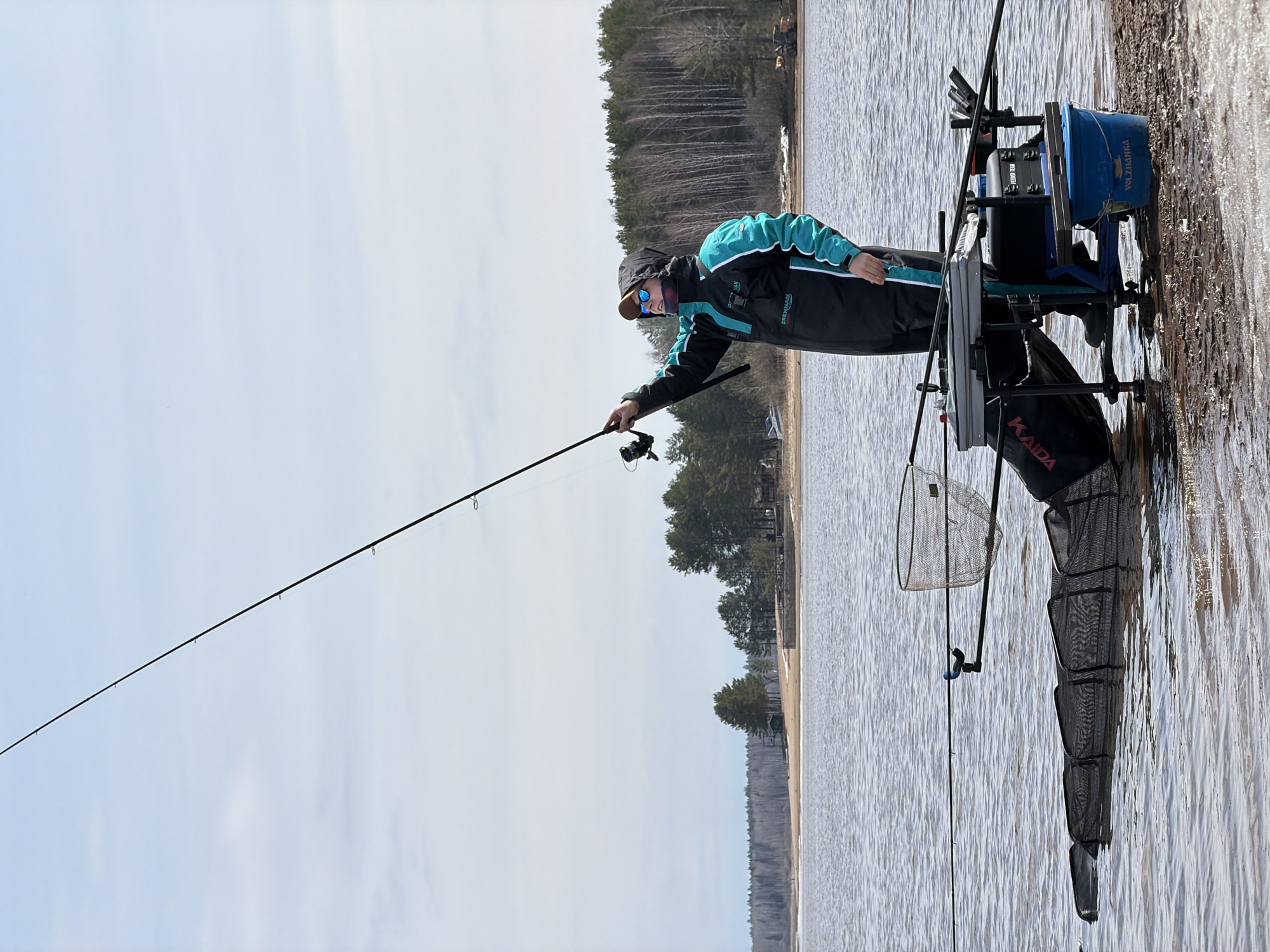 An outdoor scene in Chaykovsky, Russia, captures a man engaged in fishing on the edge of a large body of water. The image shows the man, dressed in a teal and black jacket with "DRENAN" visible, a cap, and sunglasses, holding a fishing rod with a reel. His posture suggests concentration on the fishing activity.
An elaborate fishing station is set up to his right, extending into the shallow water. It features a padded seat, multiple rod holders, and various attachments. A prominent blue bucket, marked with "VOLZHAN" (likely part of "VOLZHANKA"), is attached to the station. Floating in the water beside the setup is a long, black keepnet, clearly branded "KAIDA," used for holding caught fish, and a smaller landing net is also visible.
The background presents a distant shoreline fringed by a dense forest, composed of both bare deciduous trees and green conifers, indicating an early spring or late autumn season. The sky above is bright and appears overcast or partly cloudy. The surface of the water shows ripples, suggesting a gentle breeze. The overall impression is one of a calm, focused fishing excursion in a natural, serene environment.