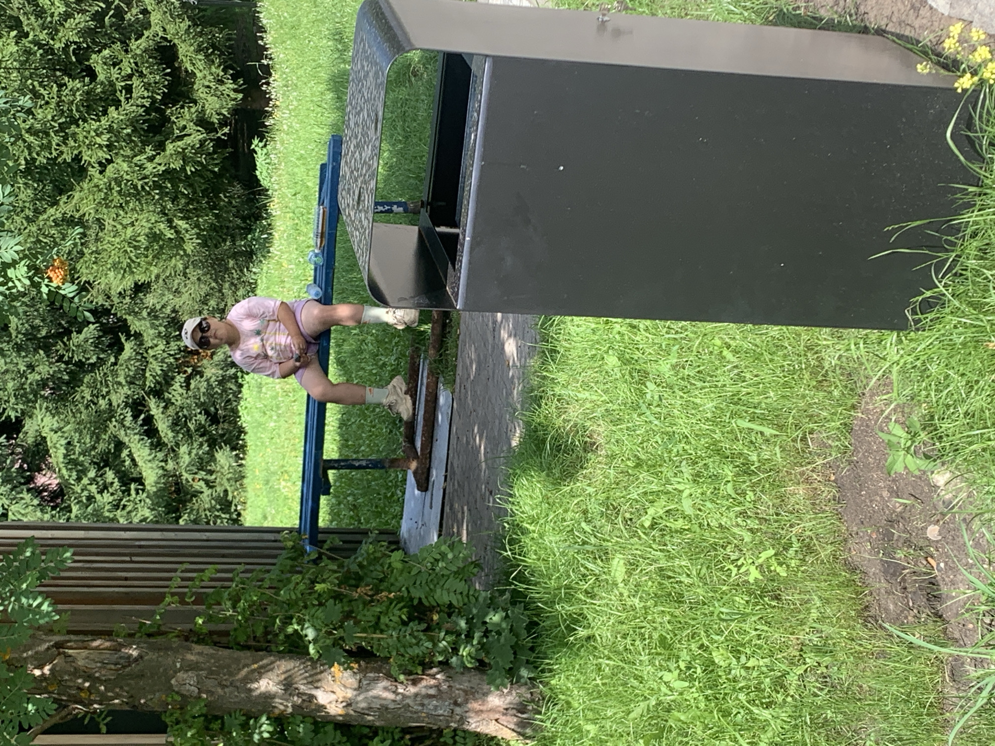 A young person wearing a pink t-shirt, shorts, sunglasses, and a cap sits on a blue park bench. The person's legs are crossed and they are holding a phone. The bench is situated in a grassy park area, with trees and foliage in the background. A black trash can is positioned in the foreground to the right of the bench. The scene appears to be in Arbuzov-Baran, Russia.