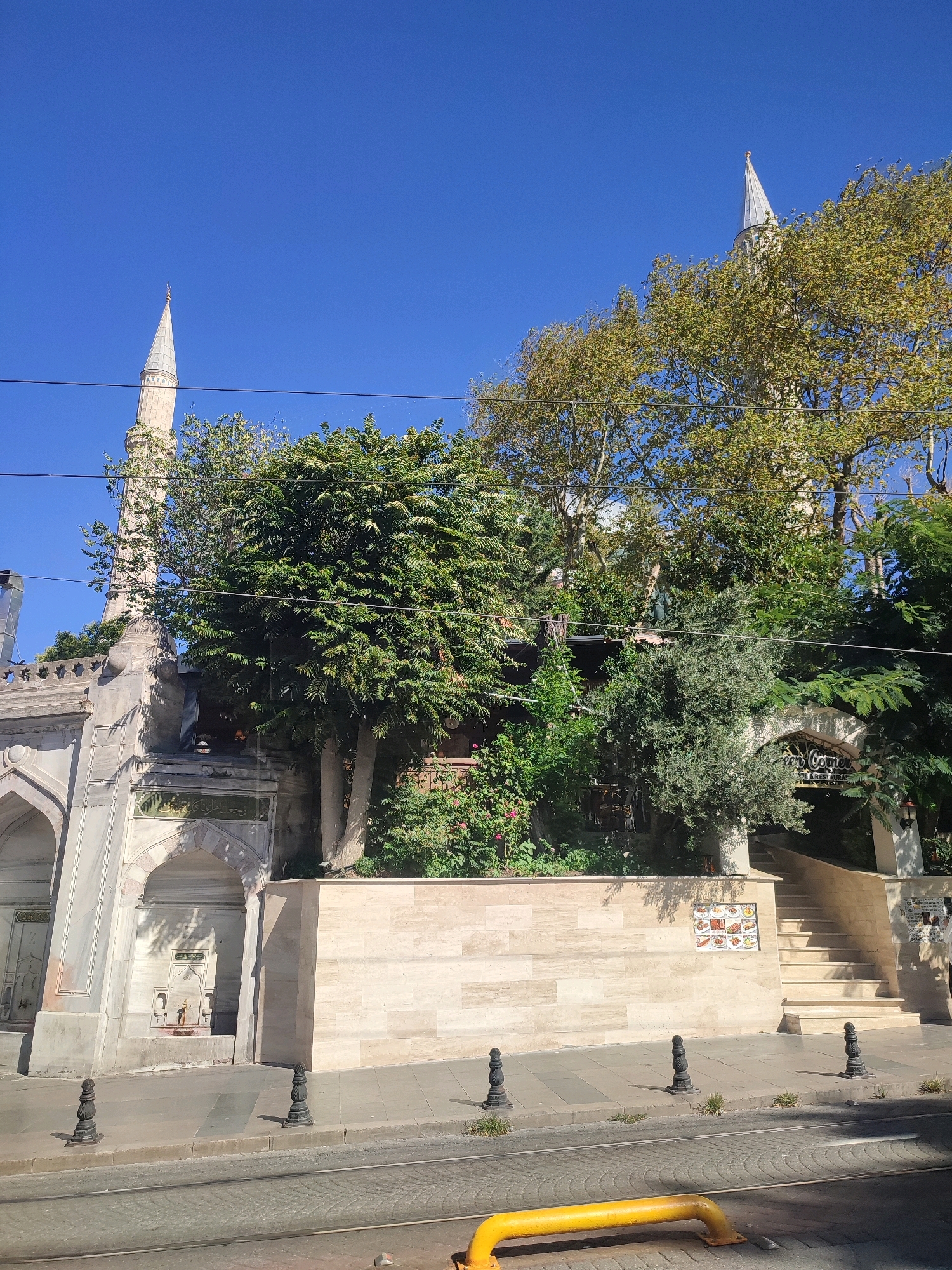 The image captures a street view in Istanbul, Turkey, featuring a mosque with two minarets. The architecture of the mosque is visible on the left, with intricate stone detailing and arched entrances. A water fountain is incorporated into the mosque's facade. Lush green trees and foliage surround the building, some of which obscure parts of the minarets. In the background, a steep set of stairs leads up to an entrance with a sign that reads "Green Corner Cafe & Restaurant". The street in the foreground appears to be a tram line, indicated by the tracks and a yellow safety barrier. The sky is clear and blue, suggesting a sunny day. There are no people visible in the frame.