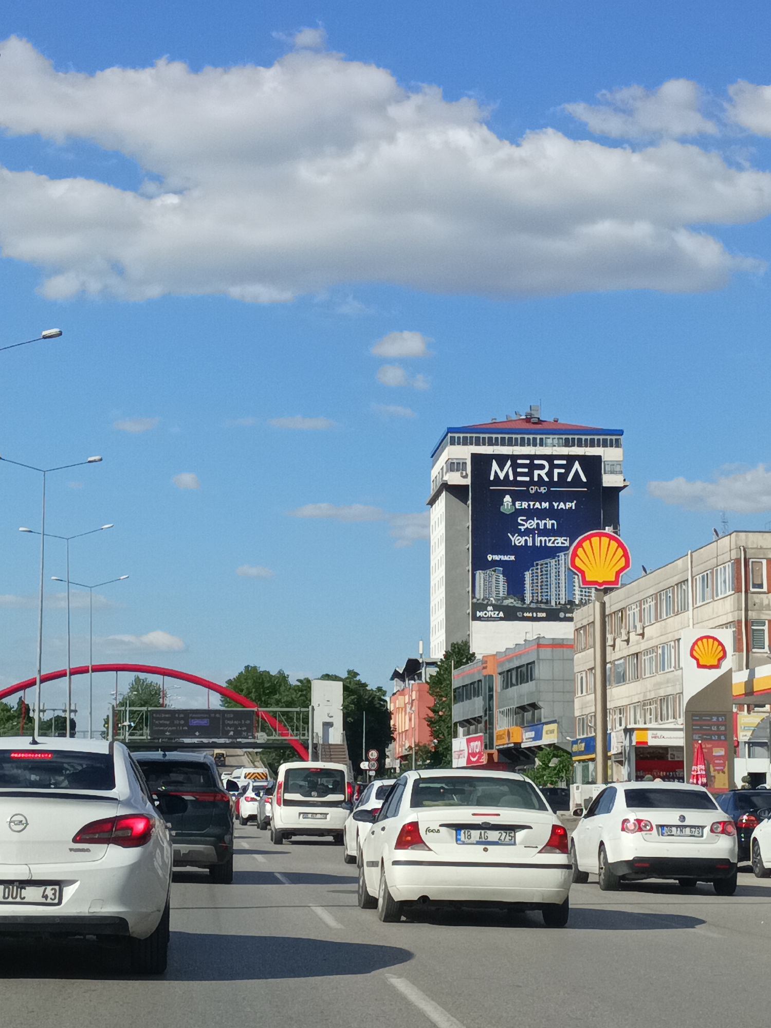 This image captures a sunny day in Altındağ, Turkey, with a clear blue sky dotted with fluffy white clouds. The scene is a bustling city street, characterized by a traffic jam on a multi-lane road. Several cars are stopped, indicating a standstill or slow-moving traffic.
In the foreground, the rear of a white Opel Astra is prominently featured, with its license plate reading "DUC 43". Other vehicles, including a dark SUV and white sedans, are visible in the traffic. A red arched bridge stretches across the road in the mid-ground, suggesting a significant architectural feature of the area.
Dominating the background is a tall, modern building with a large billboard displaying the text "MERFA grup ERTAM YAPI Şehrin Yeni İmzası". The billboard also includes architectural renderings and contact information, including "MONZA 444 8 200" and "YAPRACIK". To the right of this building, a Shell gas station is identifiable by its iconic yellow and red shell logo, with pumps and advertising visible. The overall setting is urban, with the presence of buildings, streetlights, and the highway indicating a busy commercial and residential area. The time of day appears to be daytime, given the bright sunlight and shadows.