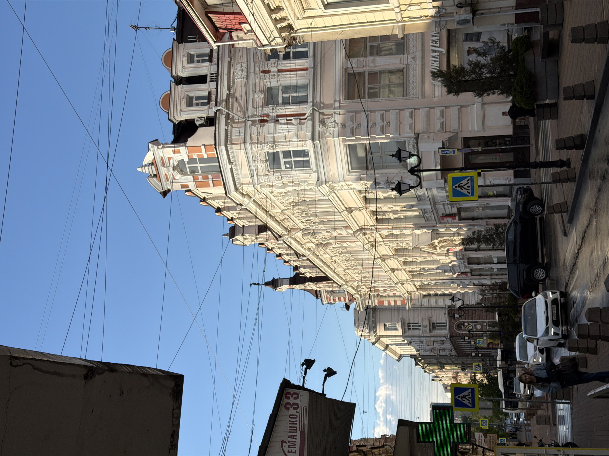 The image shows a street view in Rostov-on-Don, Russia. A multi-story building with ornate architectural details dominates the right side of the frame. Utility lines crisscross the sky. Street signs and street lamps are visible. Several cars are parked along the street. A pedestrian walks on the sidewalk. The building with the sign "ЕМАШКО, 33" is visible on the left side of the frame. The sky is clear and blue.