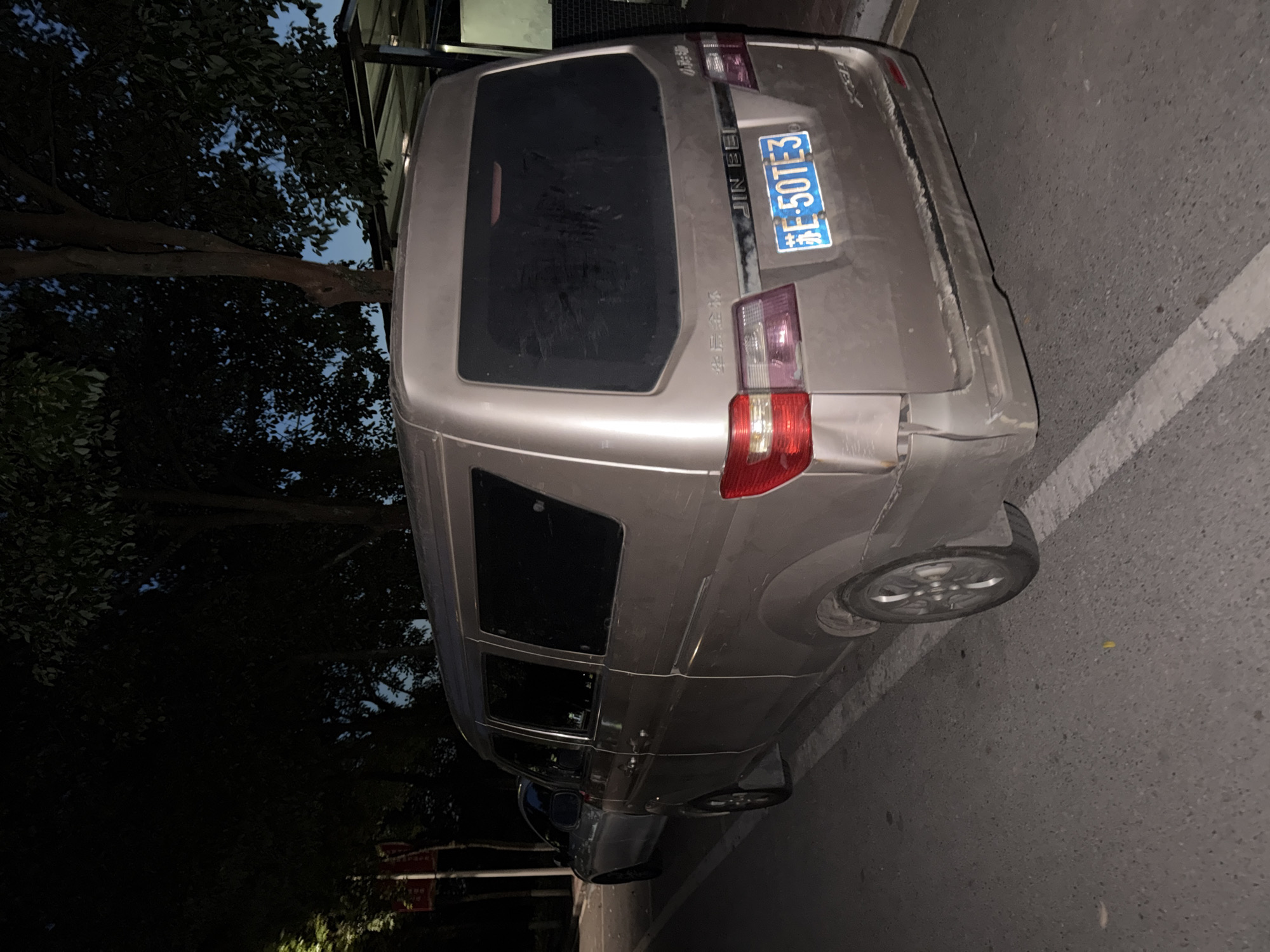 A light brown minivan is parked on the side of a road at night, with its rear facing the camera. The minivan has tinted windows and a license plate that reads "E 50TE3". There is a dark-colored sedan parked in front of the minivan, also facing away from the camera. The scene is set against a backdrop of trees and a dark sky, with a street light visible in the distance. The road appears to be asphalt and is marked with white lines. The van has some damage to its rear bumper. Based on the license plate format, the location is likely the city of Jiangjin, China.