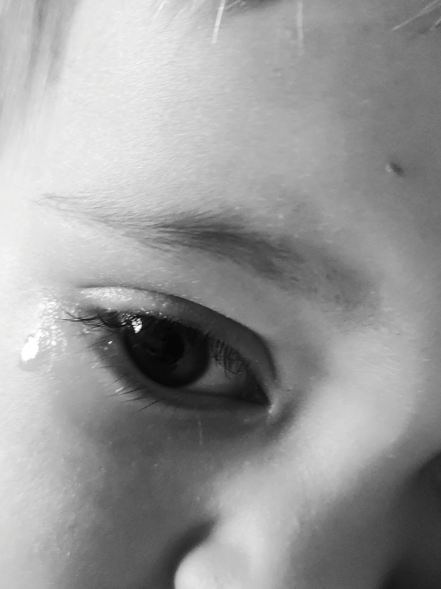 A close-up black and white photograph depicts a person's eye, eyebrow, and part of their nose. A single tear is visible below the eye. The environment is indeterminate. Location: Unknown, Unknown.
