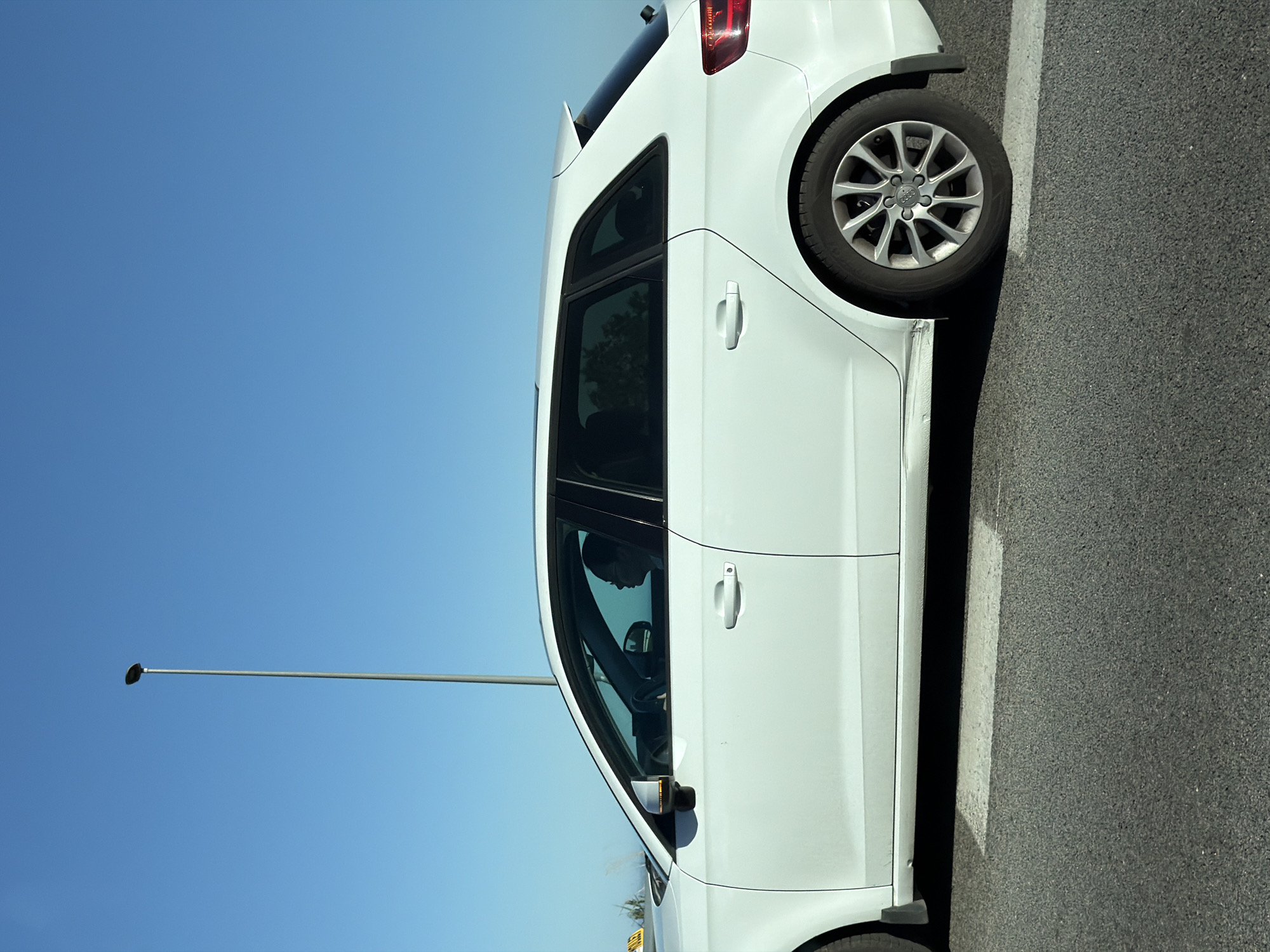 The image shows a white Audi A3 parked on the side of a road. The car is positioned on the right side of the image, and the clear blue sky is visible in the background. A lamppost is seen in the left side of the picture. The vehicle is parked next to a white line on the asphalt road. The setting of Mataró, Spain, is not definitively evidenced from the photo alone.
