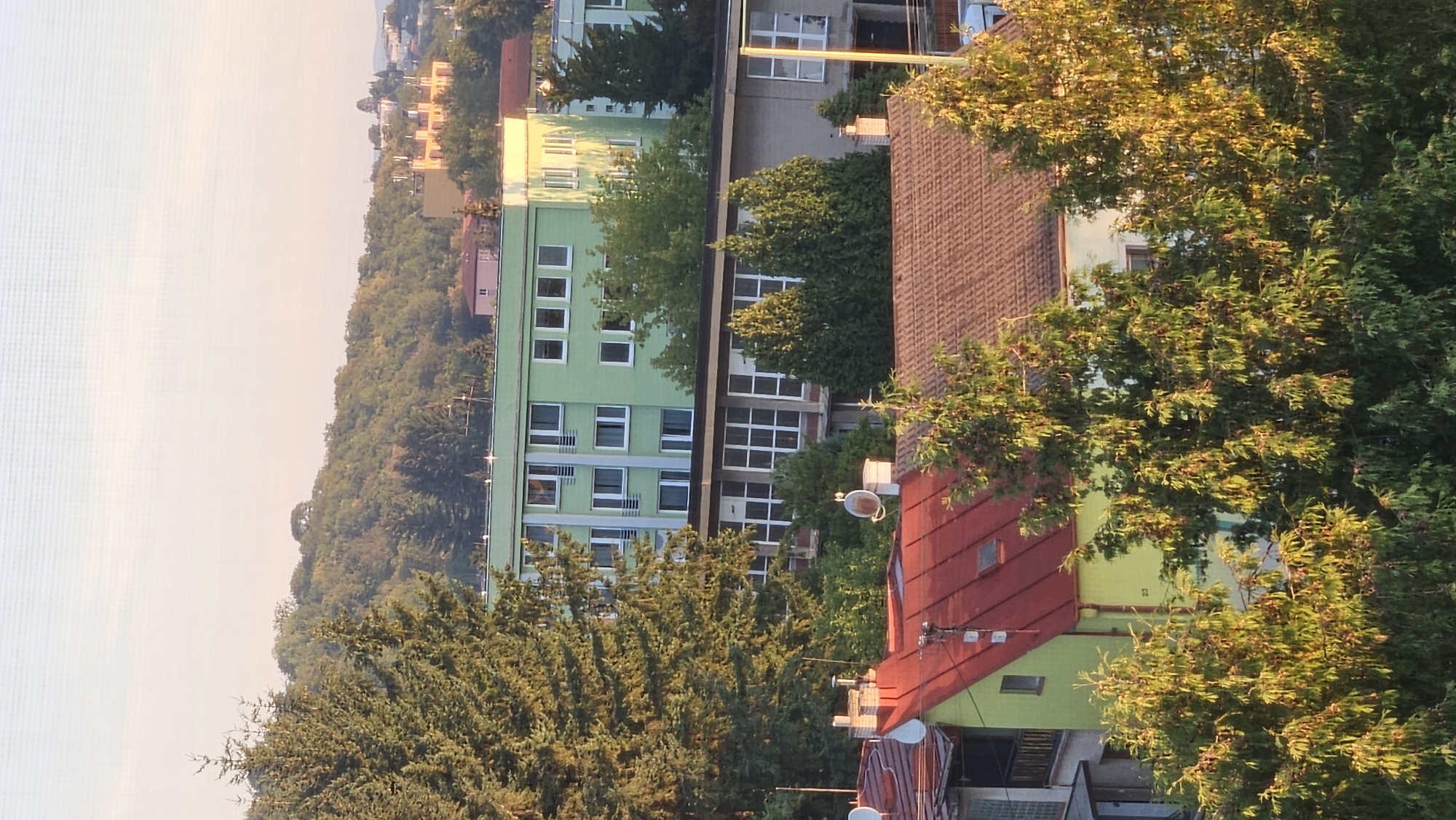 The image shows a view of a residential area in Prešov, Slovakia, with several buildings and trees. In the foreground, there are two houses with red and yellow roofs, surrounded by green trees. Behind them, there is a multi-story green building, and further in the background, more buildings and a tree-covered hill. The sky is clear and pale, suggesting it is either early morning or late afternoon. There are no people or animals visible in the image. The buildings appear to be of modern construction. The scene is peaceful and suburban.