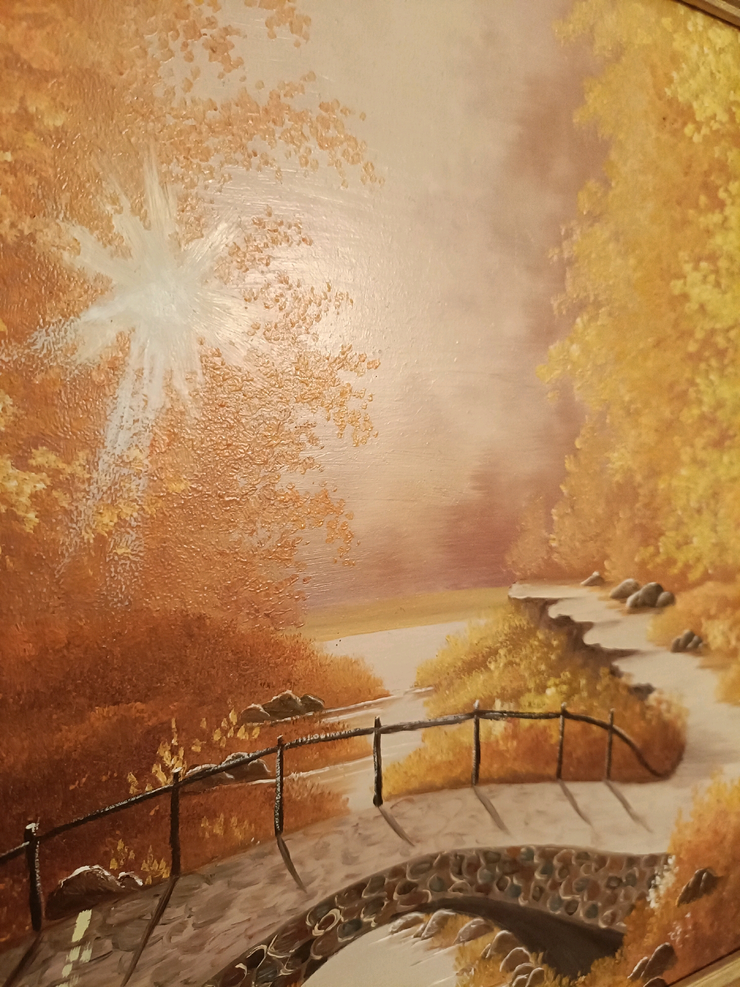 This is an oil painting of a landscape scene, possibly in autumn, featuring a stone bridge arching over a stream or path. The bridge is made of rounded stones of various sizes and colors, and it has a dark, rustic railing. A path or road, also appearing to be paved with stones or gravel, curves away from the bridge and into the background.
The surrounding environment is densely populated with trees and foliage, depicted in warm tones of orange, yellow, and brown, suggesting fall colors. Sunlight streams through the trees from the upper left, creating a bright, almost starburst effect and casting shadows on the path. The overall atmosphere is serene and natural. There are no people visible in the painting. The painting does not depict any specific location in the city of Tabuaço, Portugal.