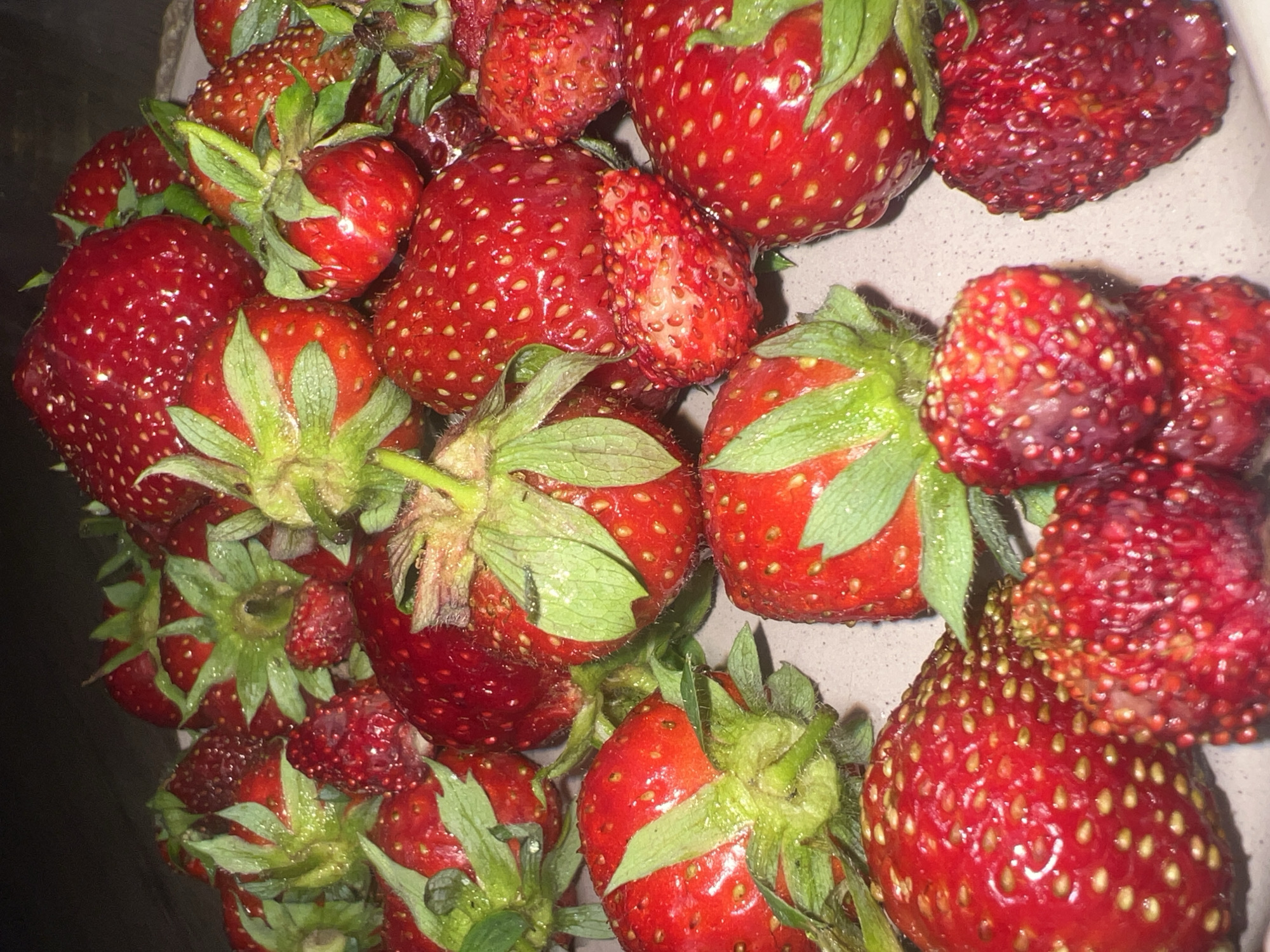 A close-up, high-angle shot showcases a pile of ripe, red strawberries, many with green leafy tops still attached. Some strawberries are large and plump, while others are smaller and more delicate. The texture of the strawberries is visible, with many tiny seeds dotting their surfaces. A few strawberries show slight imperfections or bruising, but most appear fresh and juicy. The background is dark and indistinct, drawing all attention to the fruit. The scene appears to be indoors, possibly on a plate or in a bowl. No information about Votkinsk, Russia can be derived from this image.