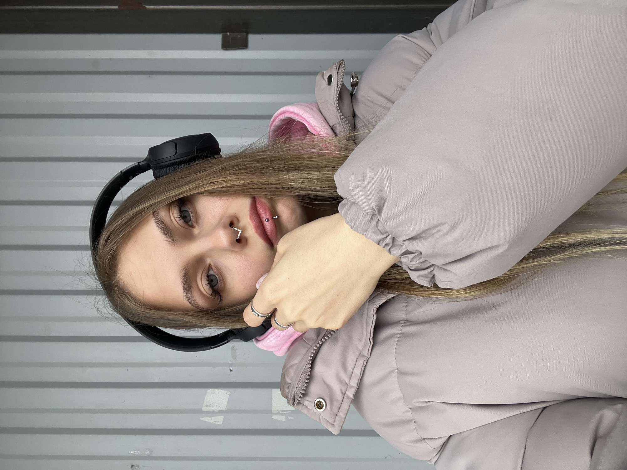 A young woman poses for a photo against a corrugated metal wall. She is wearing headphones, a pink hoodie, and a beige puffer jacket. She has facial piercings on her nose and lip. The location is in Фоки, Russia.