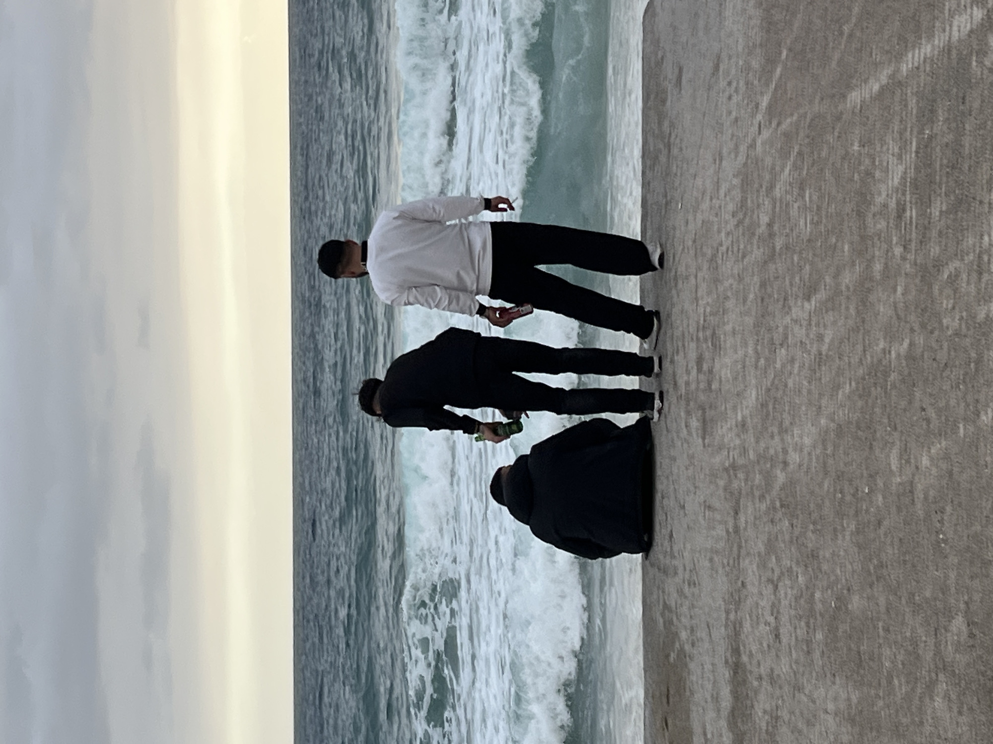 Three individuals stand on a concrete pier overlooking a turbulent sea. The pier runs horizontally across the lower half of the image. Waves crash against the pier's edge. The sky is overcast and greyish.
The first individual, closest to the top of the image, wears a white sweatshirt and dark pants. He holds a cigarette in his right hand and a mobile phone in his left. The second person, below him, is dressed in a black jacket and dark pants, holding a green bottle in his left hand. The third person, at the bottom, is wearing a black jacket. The individuals are facing toward the sea, near Alanya, Turkey.