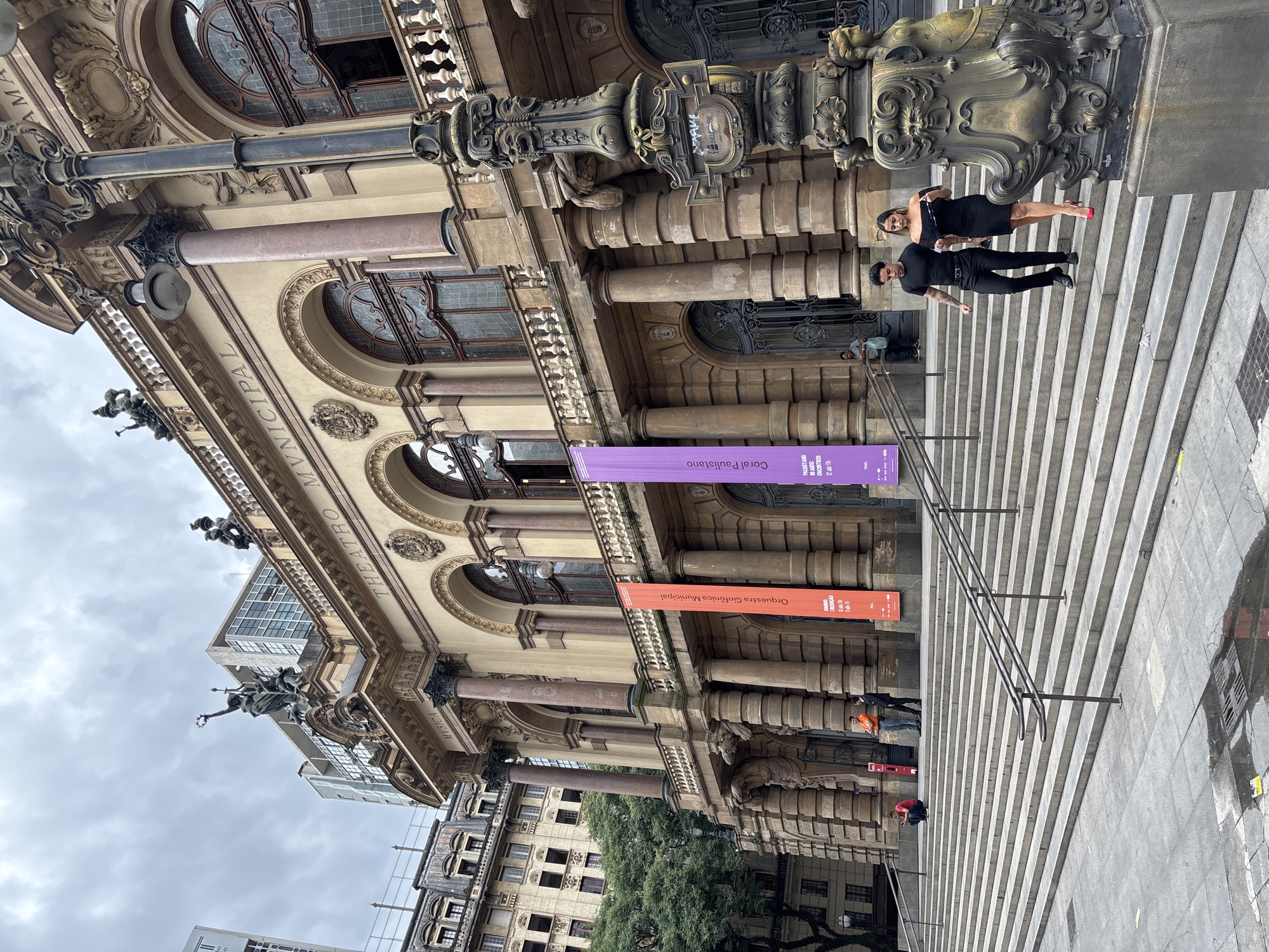 The image captures the facade of the Theatro Municipal de São Paulo, Brazil, a historical building with ornate architectural details, including pillars, archways, and sculptures. Two banners are hung on the facade, one purple and one orange, displaying information about upcoming events or performances. A man and a woman are on the staircase of the theater. The weather appears to be overcast. The scene is a public space in the city, with people visible on the steps.