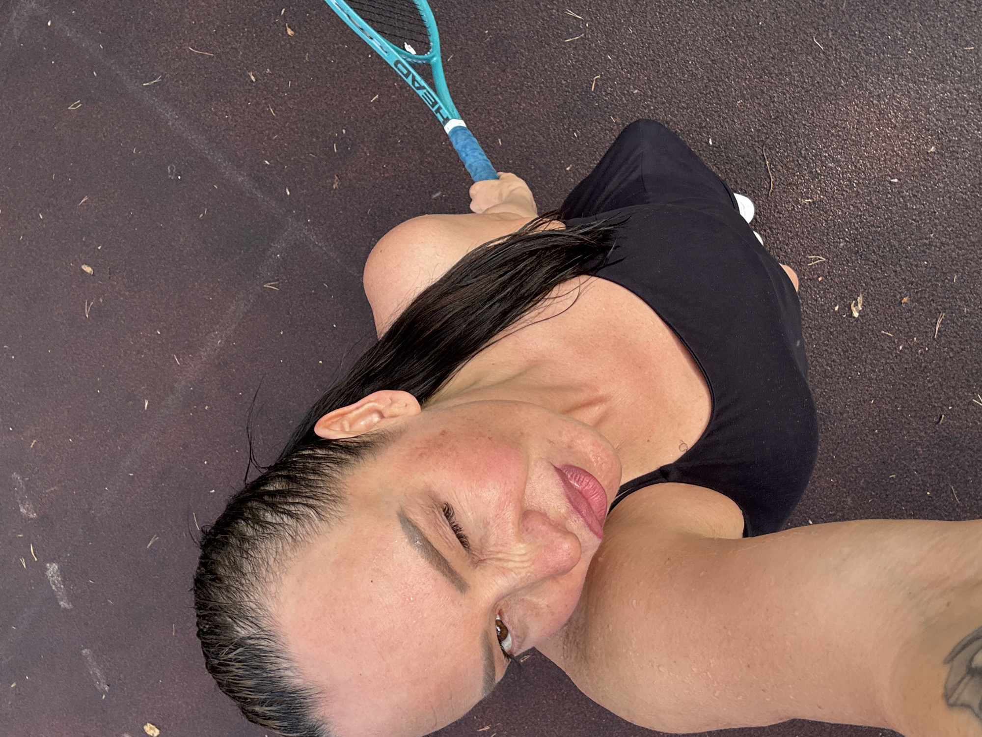 A top-down selfie shows a woman with wet, dark hair, wearing a black top, and holding a blue Head tennis racket. She is standing on a dark, textured surface that appears to be a tennis court. The location is identified as the city of Прикамский, Russia. The woman's face shows signs of perspiration.