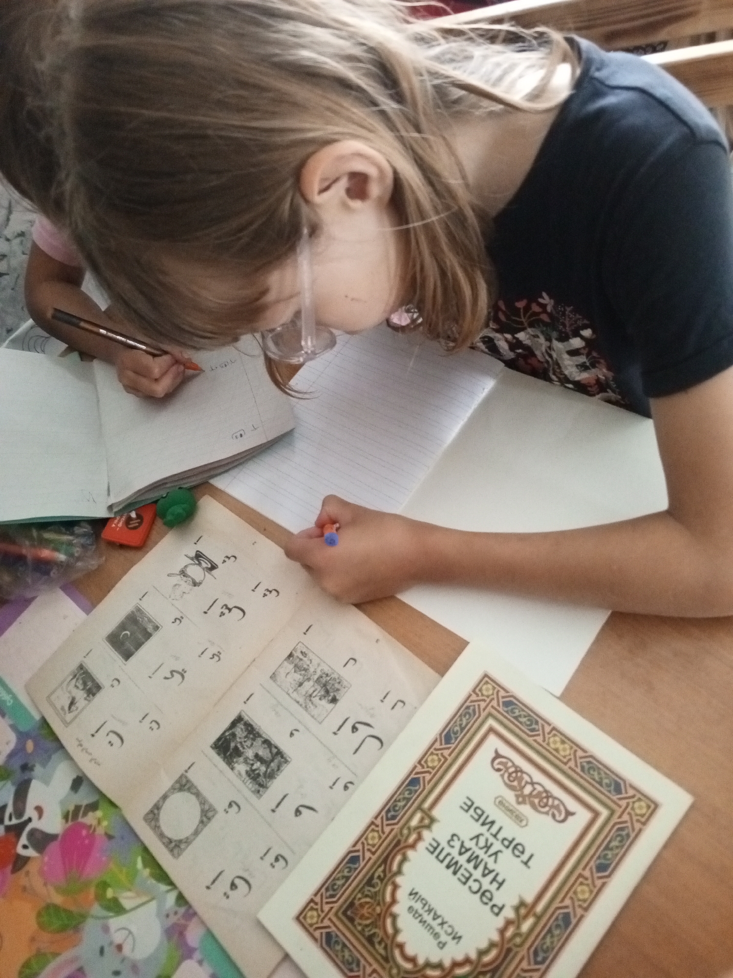 The image shows a young person with blonde hair, wearing glasses and a dark blue t-shirt with a floral print, leaning over a table and writing in a notebook. The person is holding a pen in their right hand and is focused on their work. There are several books and papers spread out on the table, some of which appear to contain text and illustrations in a foreign language, possibly Arabic or a related script. A red eraser and a green object, possibly a pencil sharpener, are also visible on the table. The environment suggests a study or learning setting, potentially at home or in a classroom. The image does not provide any specific visual cues to determine its exact location within the city of Buraly, Russia, or any other geographical context.