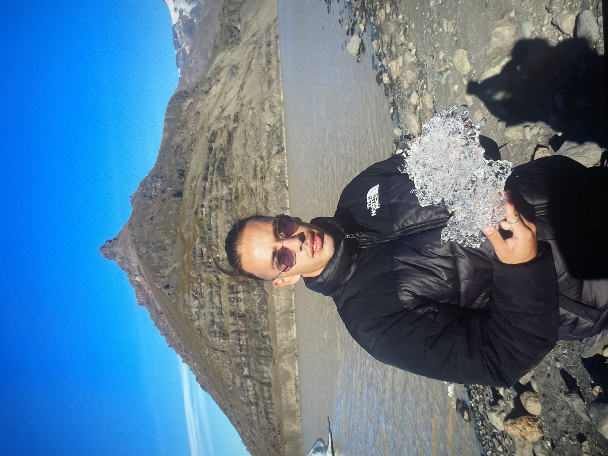 A medium shot captures a man in sunglasses and a black puffer jacket holding a chunk of ice. He is positioned in front of a body of water with a rocky shoreline. In the background, a large mountain rises to the left of the frame under a clear blue sky. The location is Unknown, Unknown.