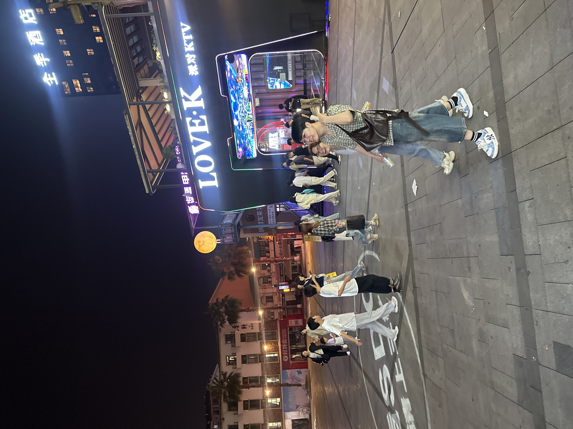 The image depicts a night scene in a bustling urban plaza, possibly within the Chongqing High-tech Zone, China. In the foreground, two individuals are walking towards the viewer, one with a checkered shirt and glasses, carrying a brown shoulder bag, and the other in denim jeans. Behind them, a group of people, including children, are walking across the plaza, which is marked with large white lettering and circular patterns on the ground. To the right, a prominent building with illuminated signage reads "LOVE K" and "KTV", suggesting an entertainment venue. The background shows multi-story buildings, palm trees, and a bright, circular yellow light fixture resembling a moon. The overall atmosphere is lively, with artificial lighting from signs and buildings illuminating the night.