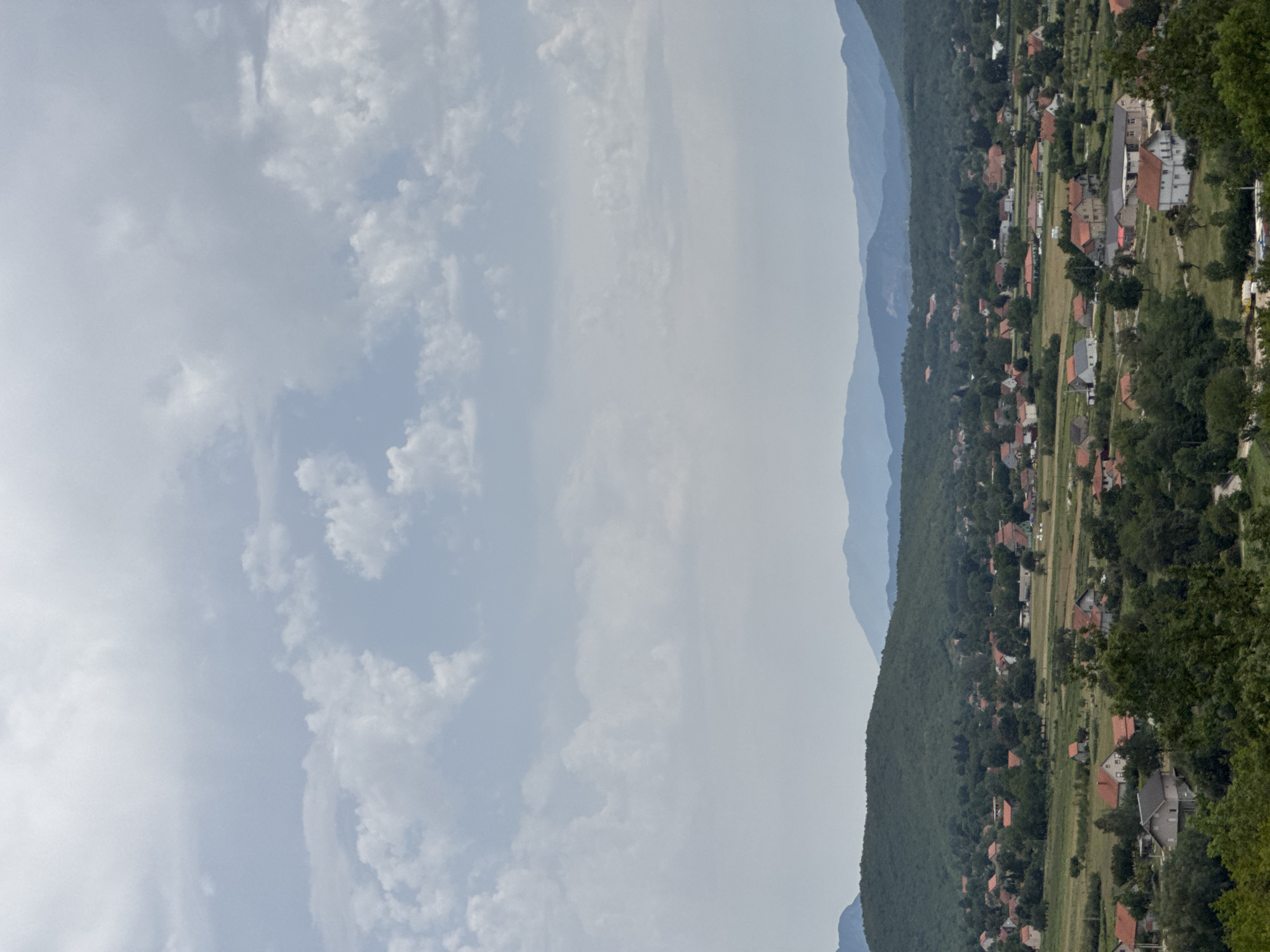 This is a wide, high-angle shot of a rural landscape in Vidrovan, Montenegro. The sky is filled with scattered white and gray clouds. In the middle ground, a village is nestled amidst green hills and fields. The village consists of numerous houses with red-tiled roofs, interspersed with trees and some open grassy areas. The houses vary in size and architectural style, with some appearing to be single-family dwellings and others larger structures. In the background, layered mountain ranges recede into the distance, their forms softened by the atmospheric haze. The overall impression is one of a peaceful, established rural community within a scenic natural environment.