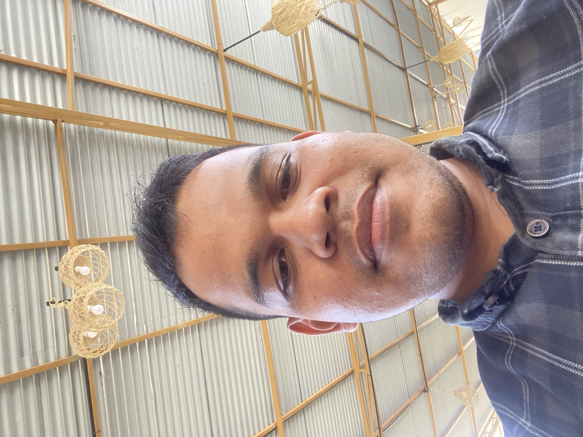 A man is shown from the chest up, looking directly at the camera in a selfie. He has dark hair and brown eyes, and is wearing a dark plaid shirt. The background is a corrugated metal ceiling supported by yellow beams. Several woven pendant light fixtures are visible hanging from the ceiling. The setting appears to be an indoor or covered outdoor space. There is no specific indication of location within Aluepiet, Indonesia.